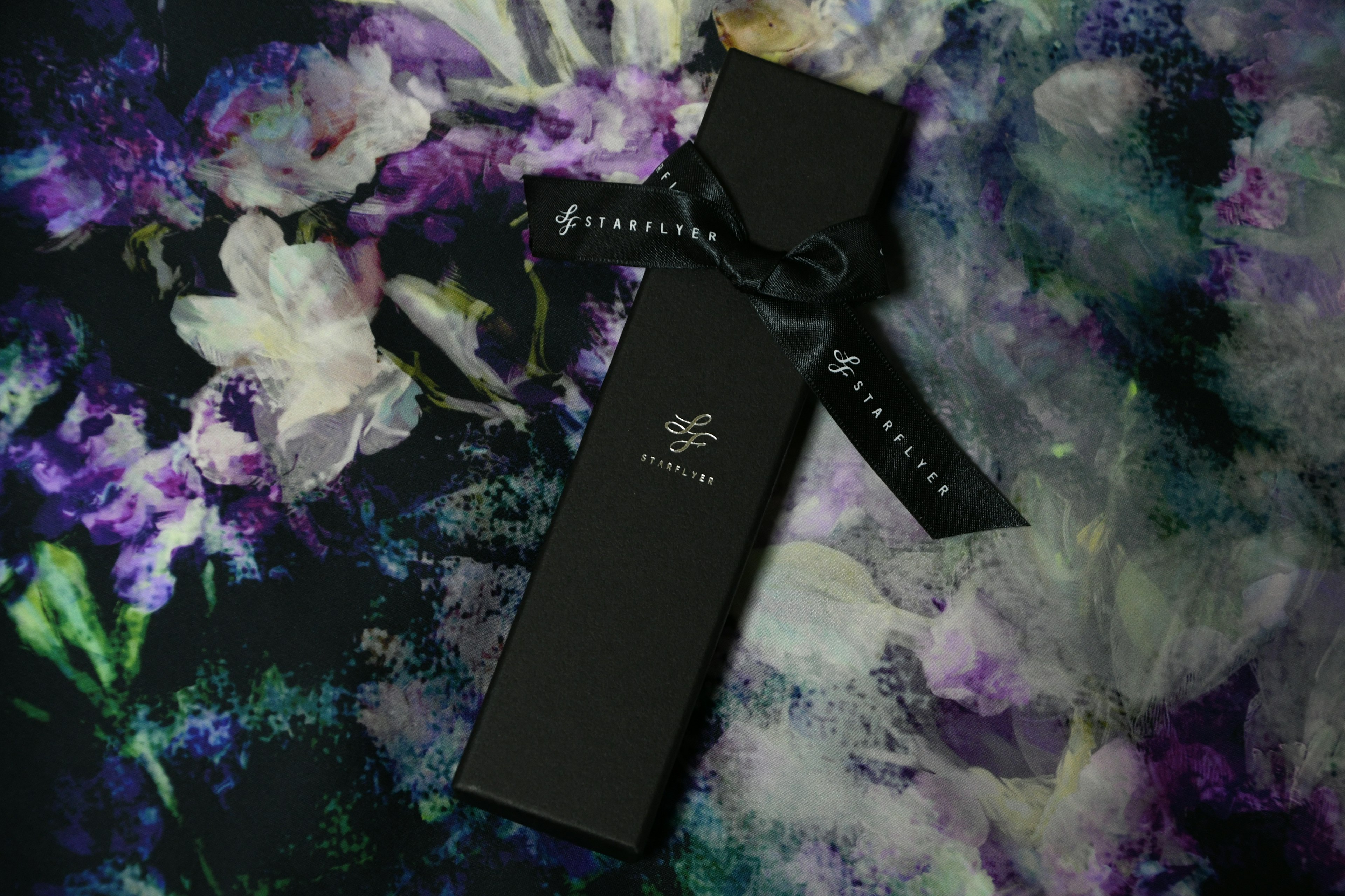 Kotak parfum hitam yang elegan dengan pita di latar belakang floral yang berwarna-warni