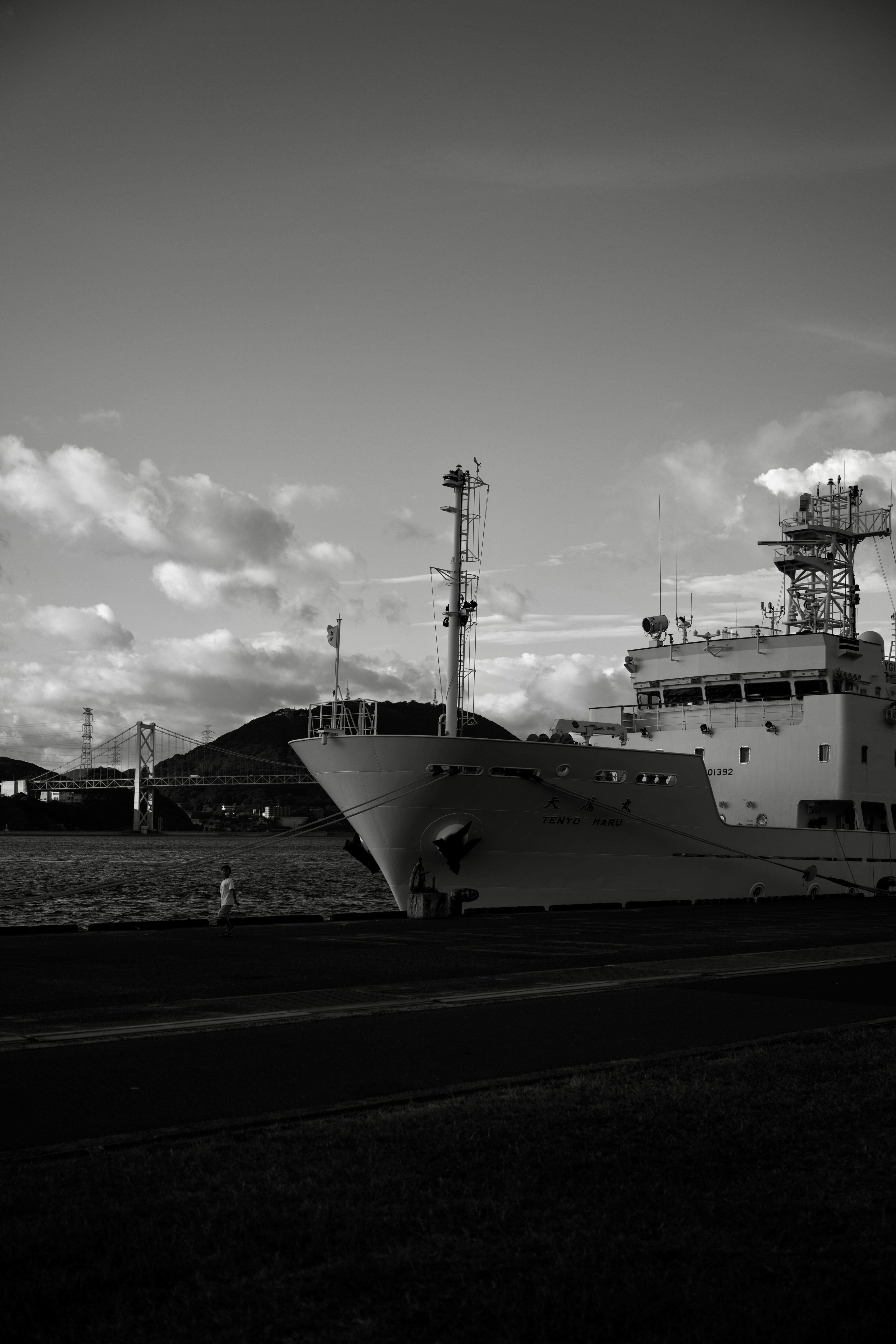 Imagen en blanco y negro de un barco con la proa a la izquierda y nubes y edificios portuarios al fondo