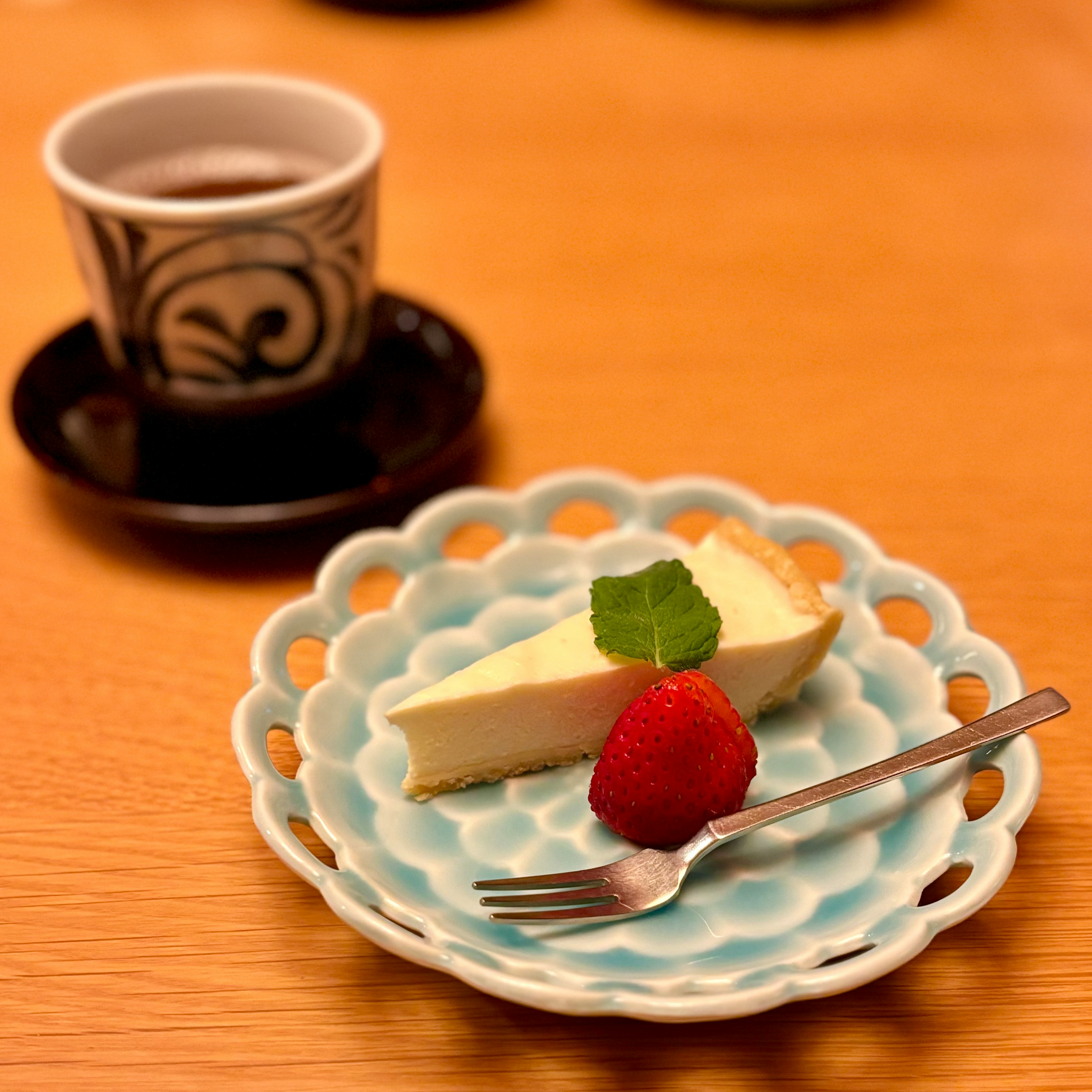 Cheesecake slice on a blue plate with a strawberry and mint leaf beside a coffee cup