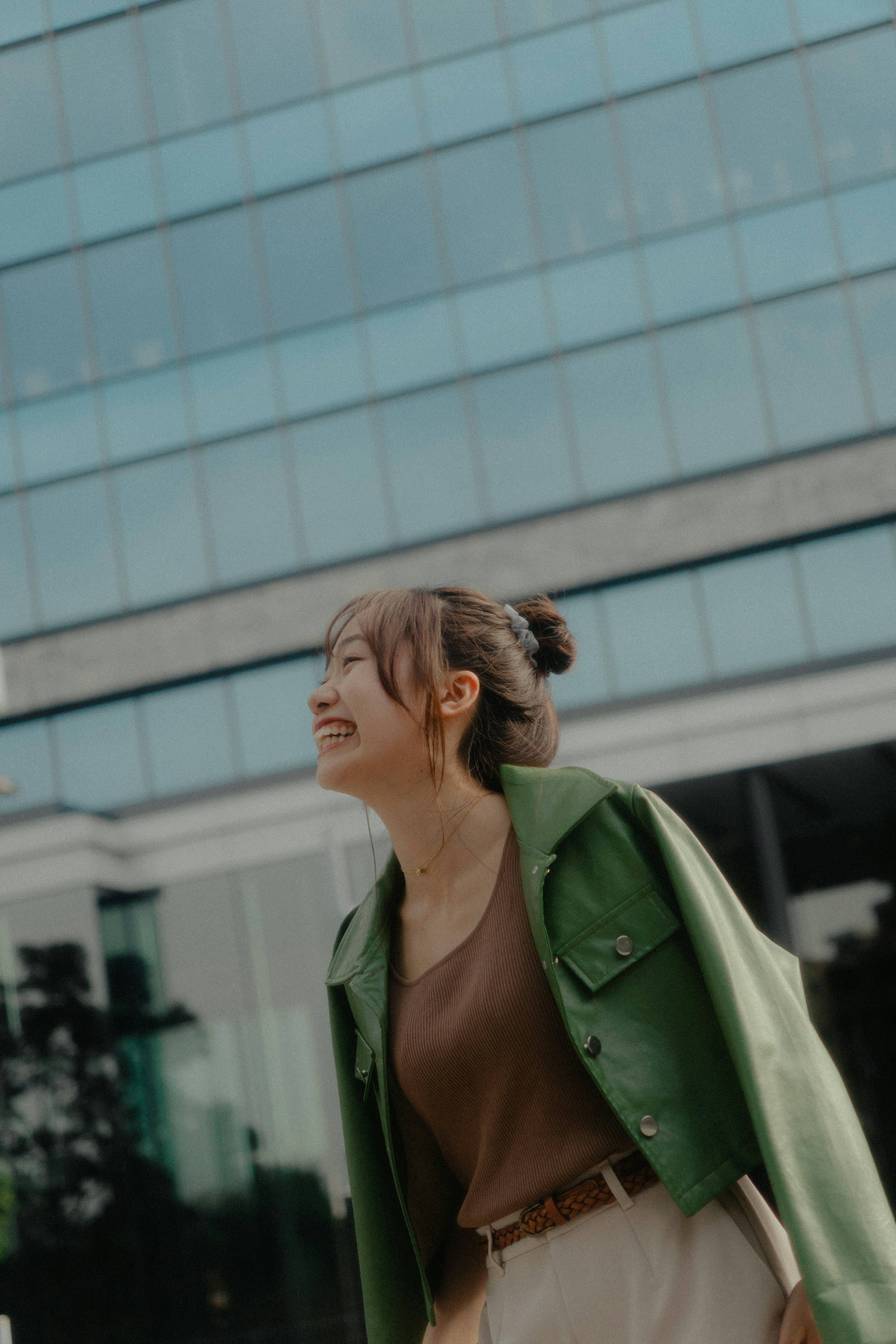 Femme en veste verte souriante à l'extérieur près d'un bâtiment en verre moderne