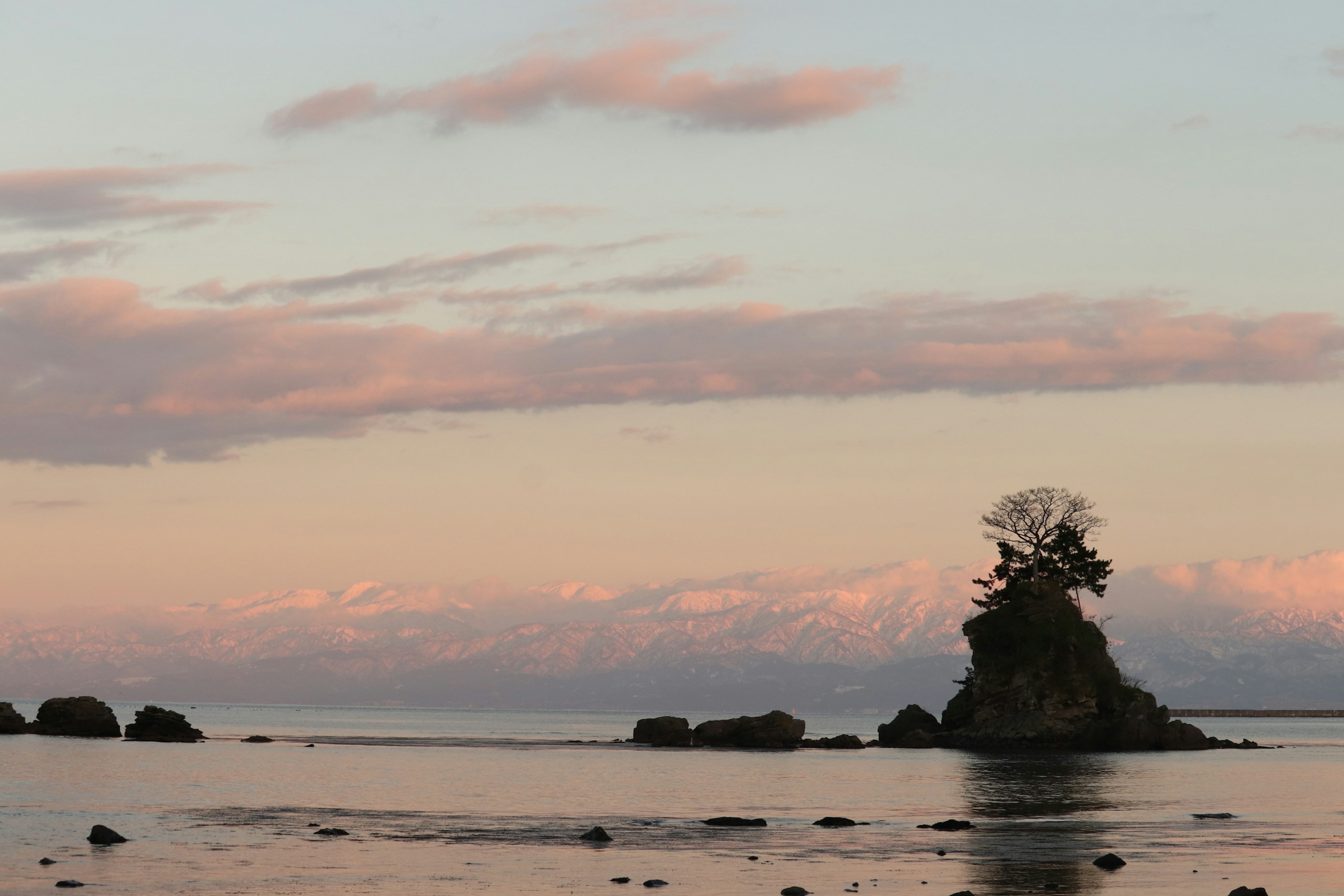 Eine kleine Insel mit Bäumen darauf in ruhigem Wasser mit schneebedeckten Bergen im Hintergrund und einem sanften Sonnenuntergangshimmel