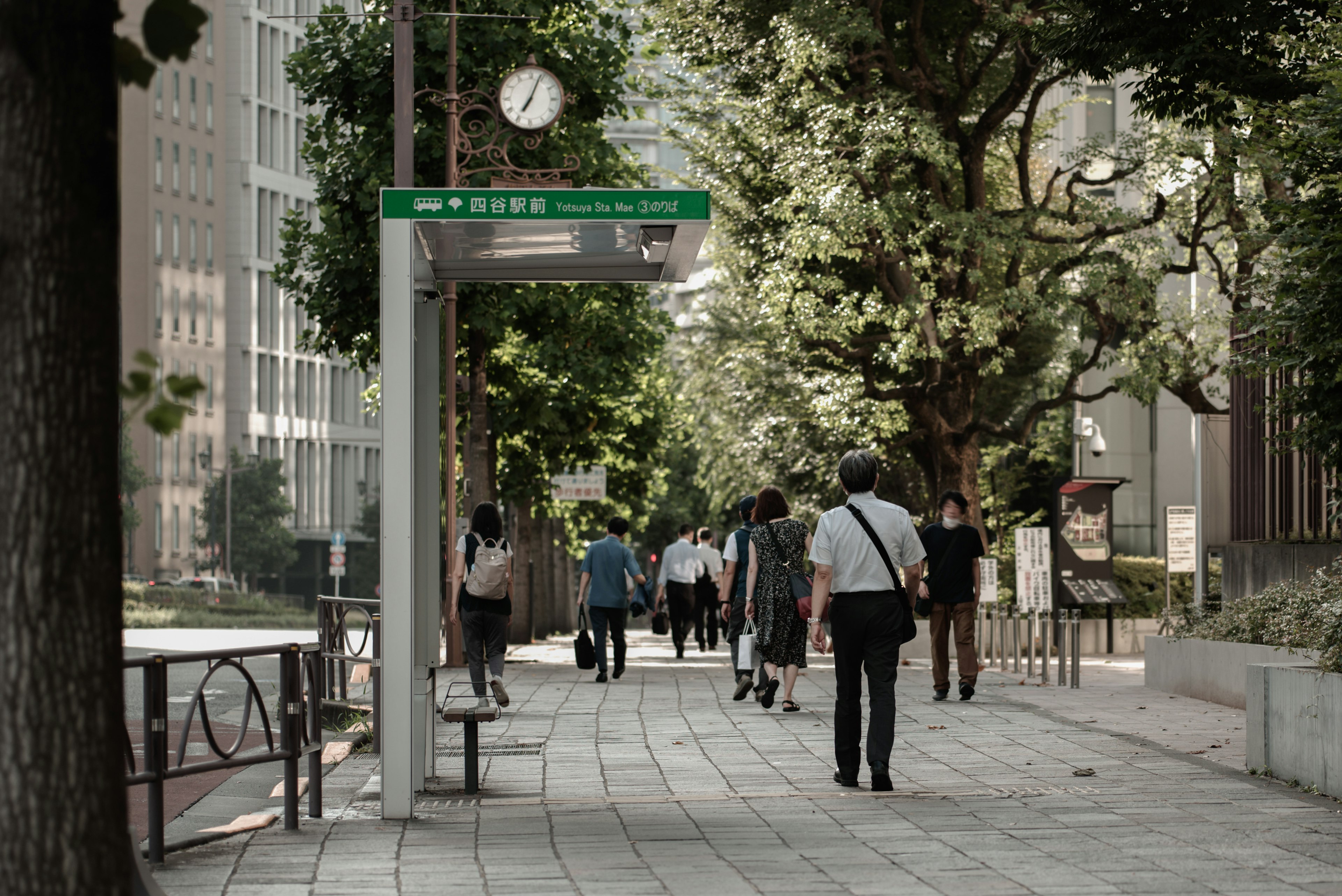 People walking along a tree-lined street with a bus stop and clock