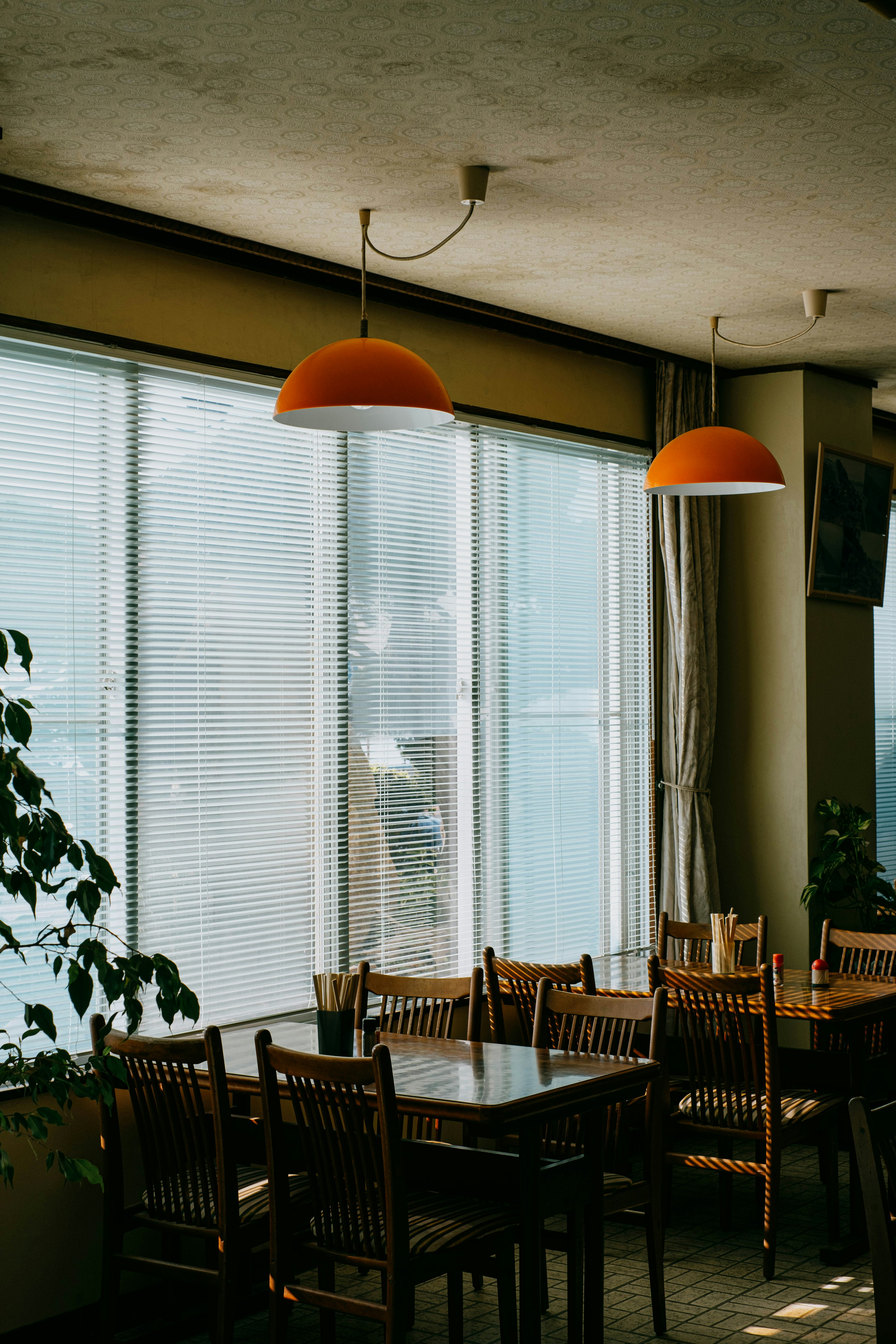 Interior de una cafetería con mesas y sillas de madera cerca de grandes ventanas luces colgantes naranjas