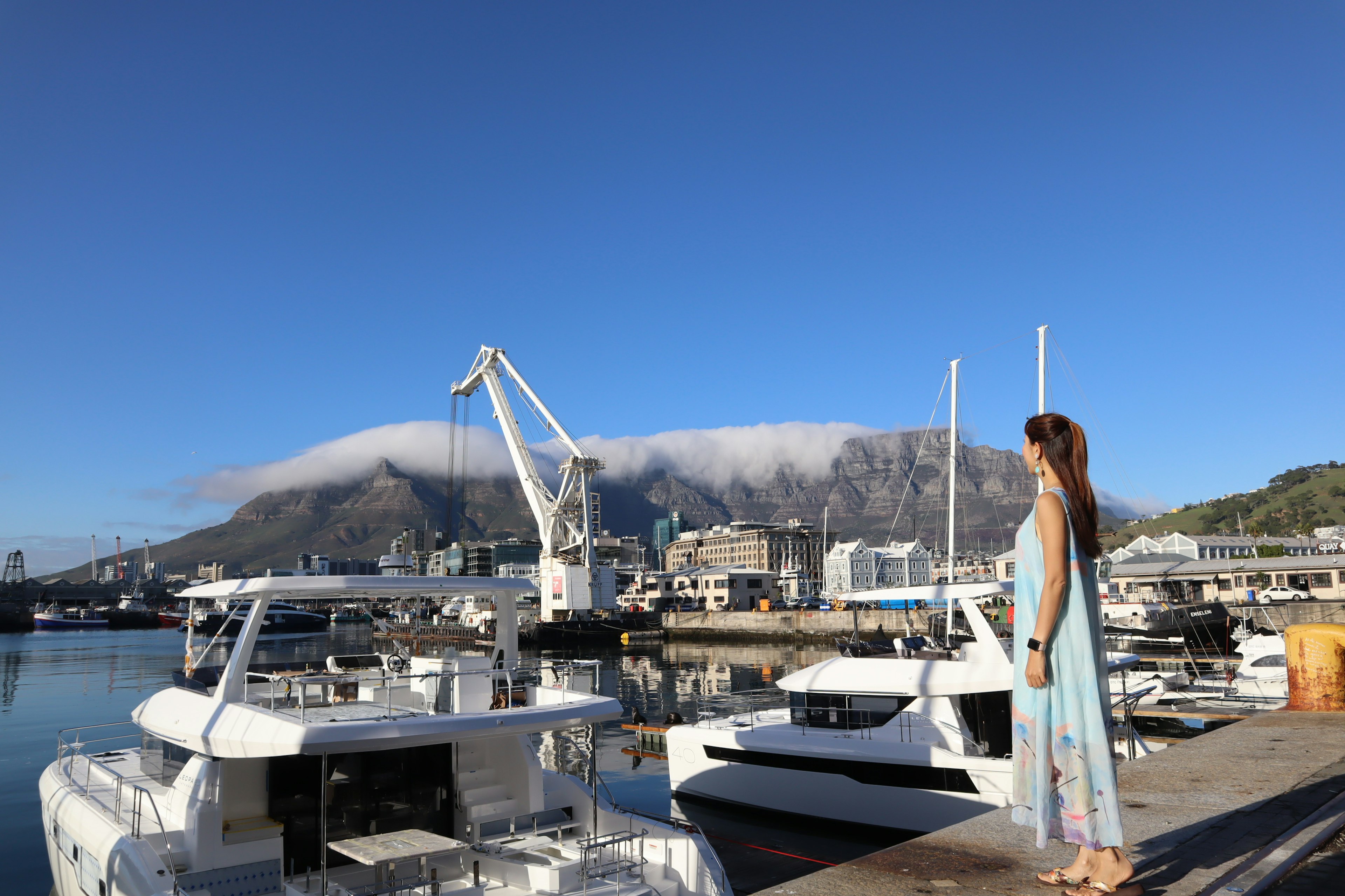 A woman standing near boats with a mountain backdrop under a clear blue sky