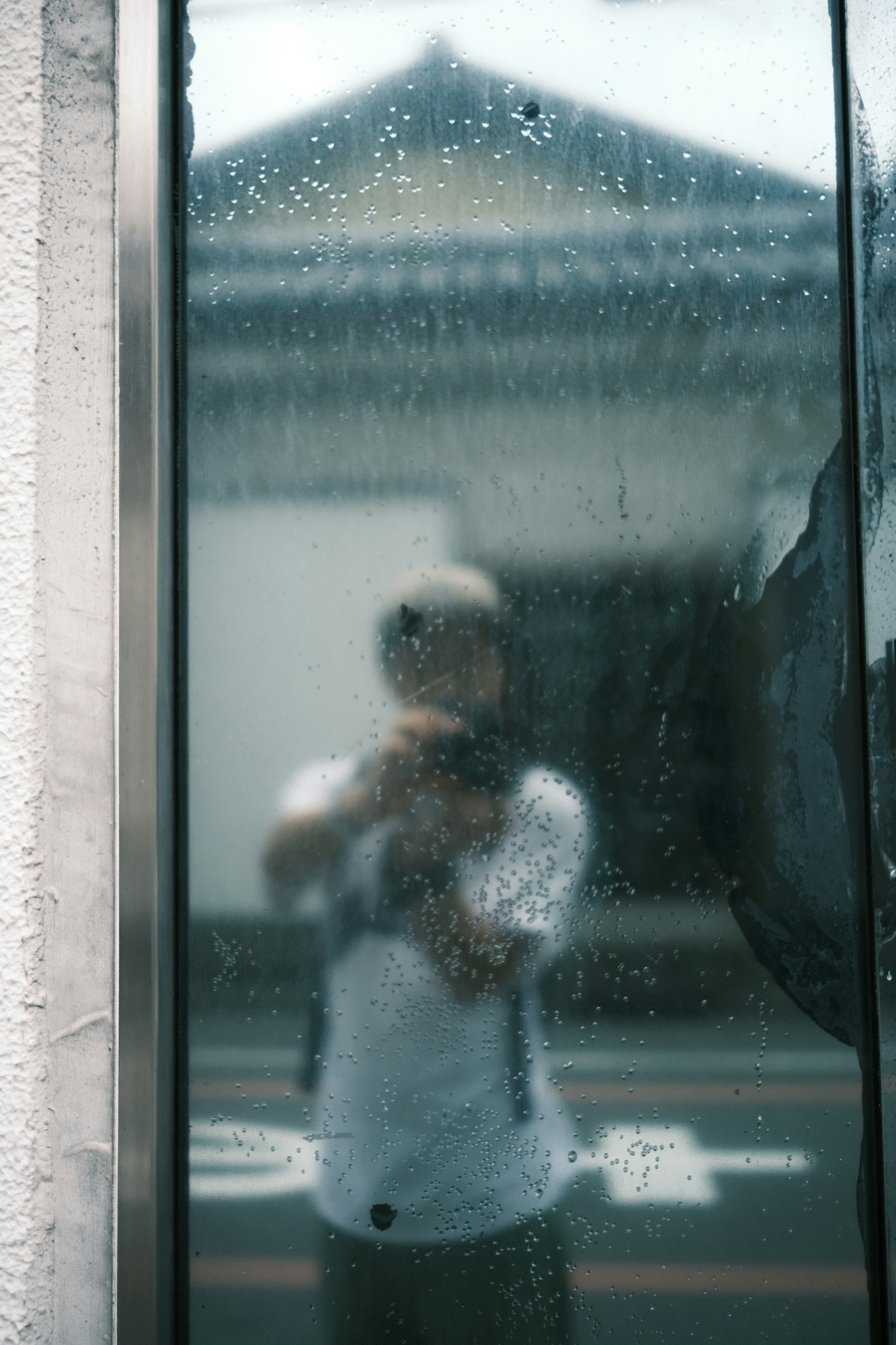 Person taking a selfie reflected in a rain-streaked glass with a traditional building in the background