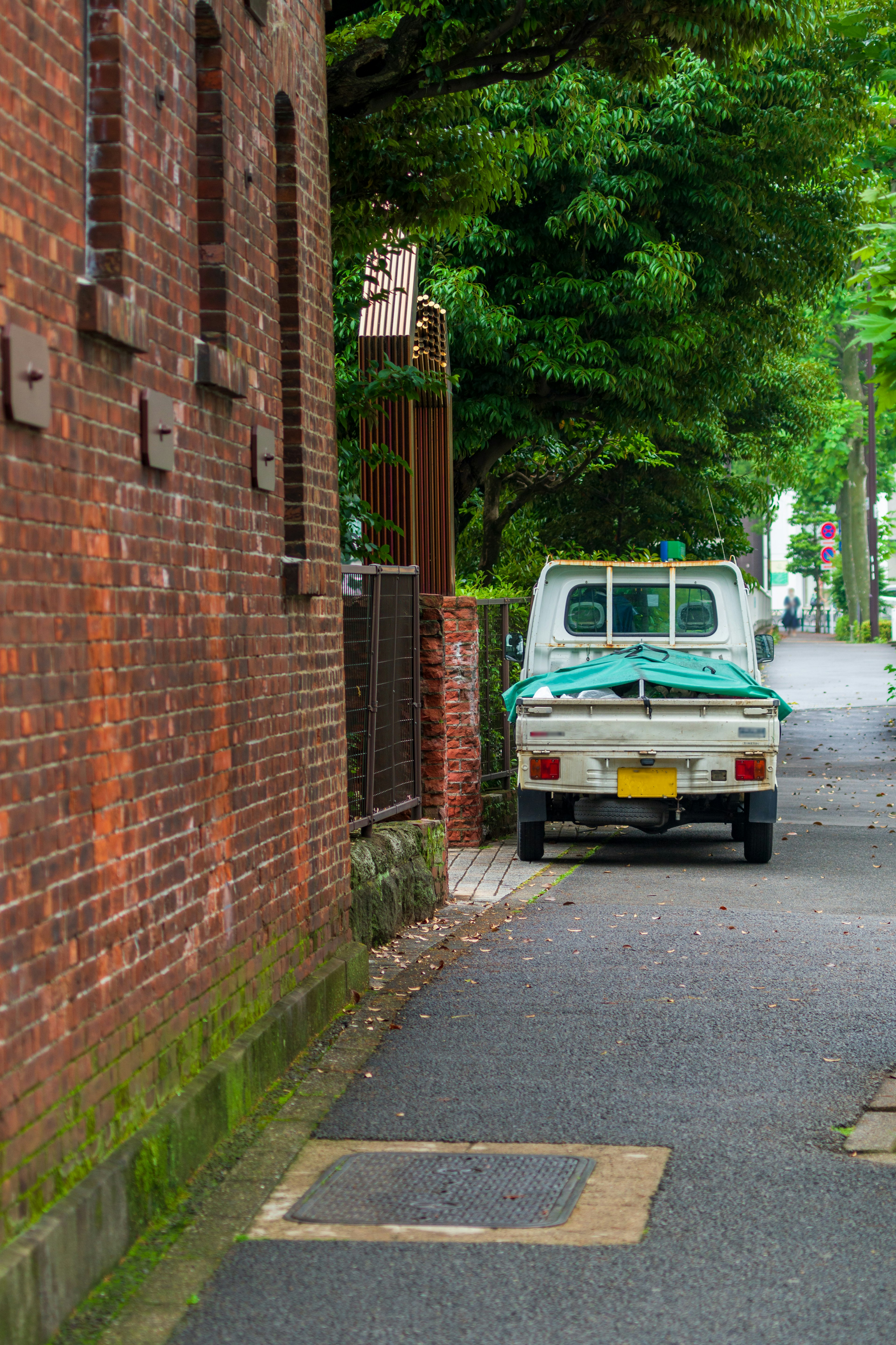 A narrow street lined with a brick wall and green trees featuring a parked light truck