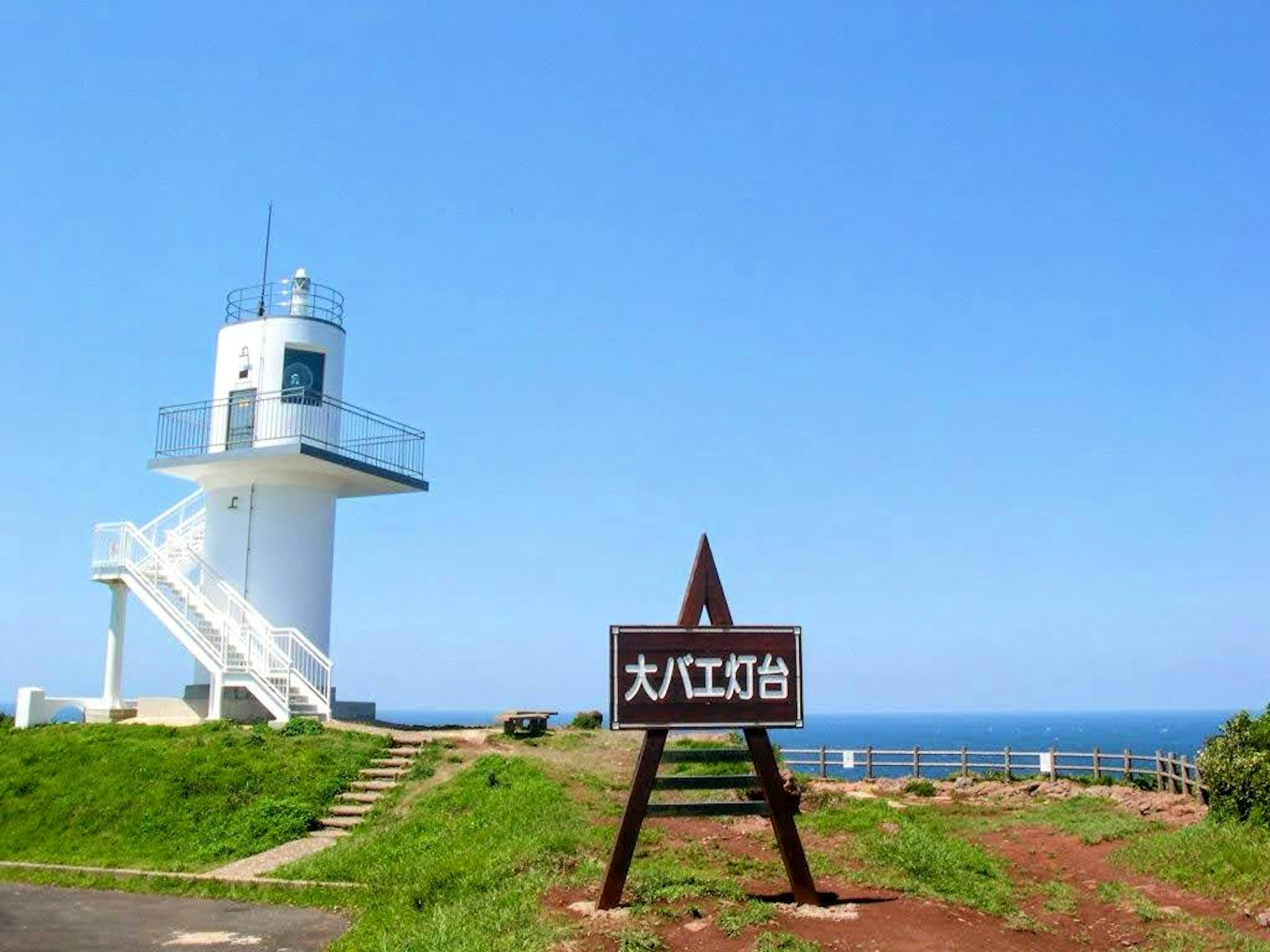 A white lighthouse beside a sign with Japanese text against a blue sky and ocean