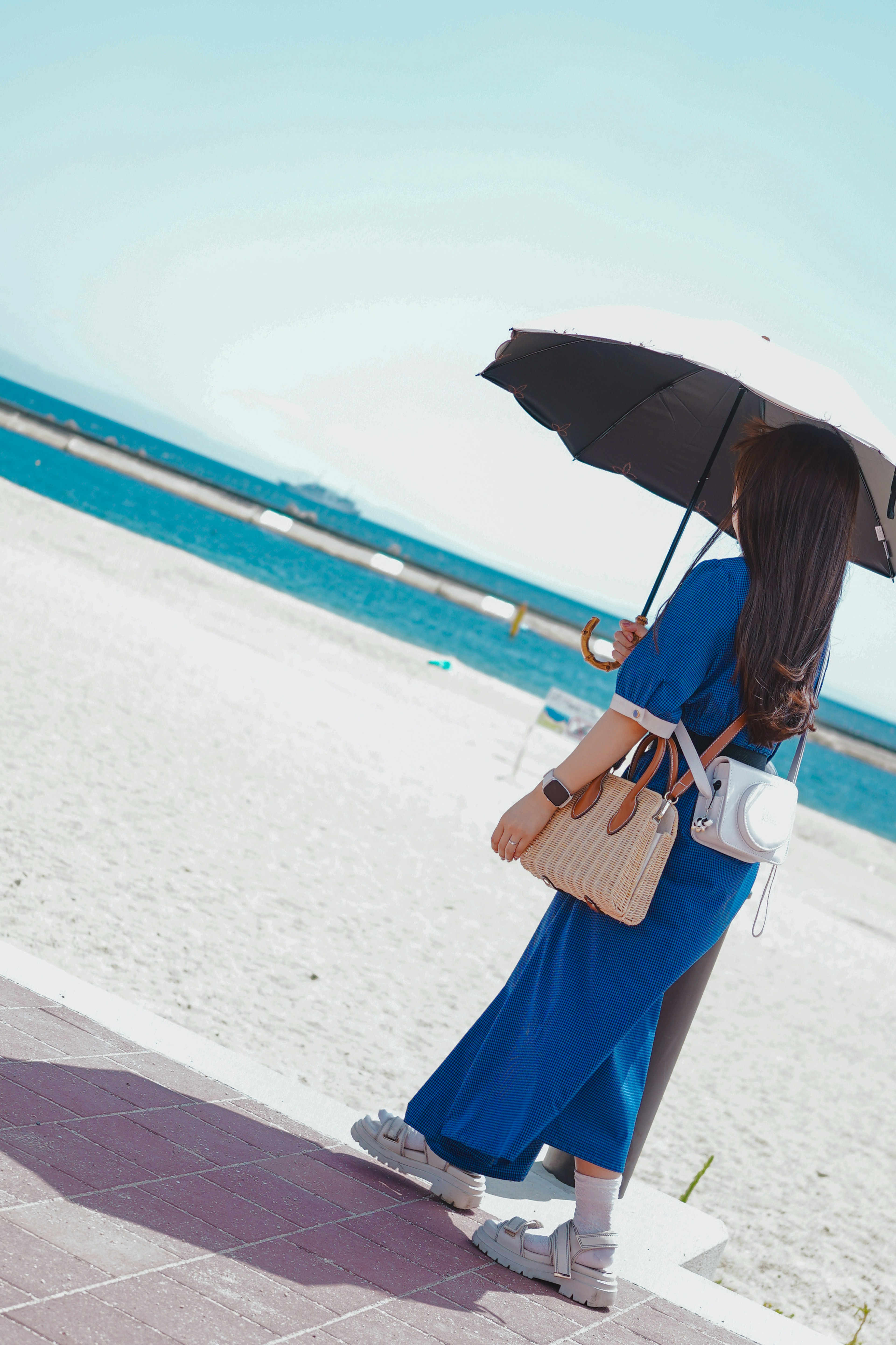 Seorang wanita berpakaian biru memegang payung berjalan di pantai