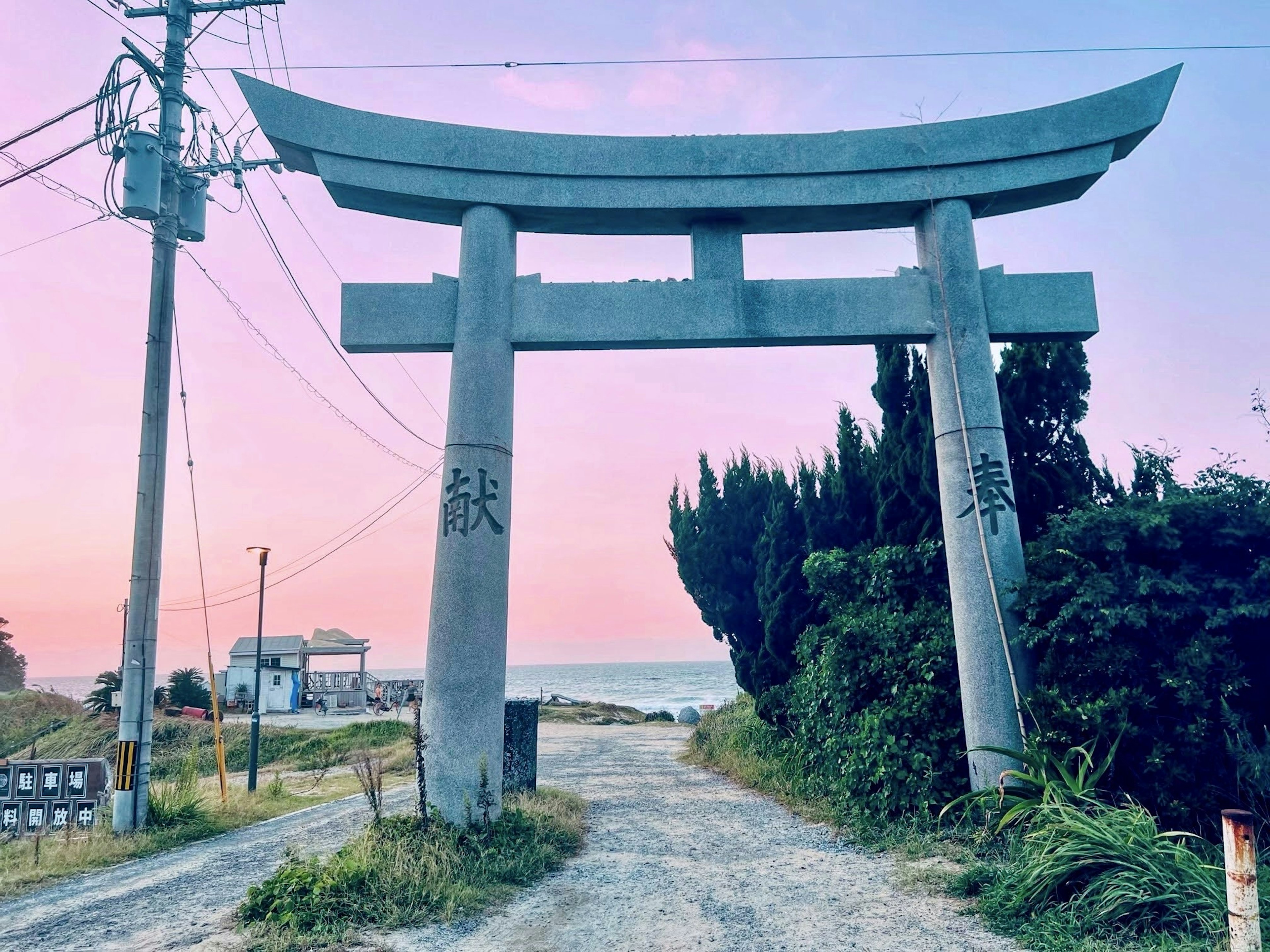 A large torii gate stands on a path leading to the sea with a sunset sky