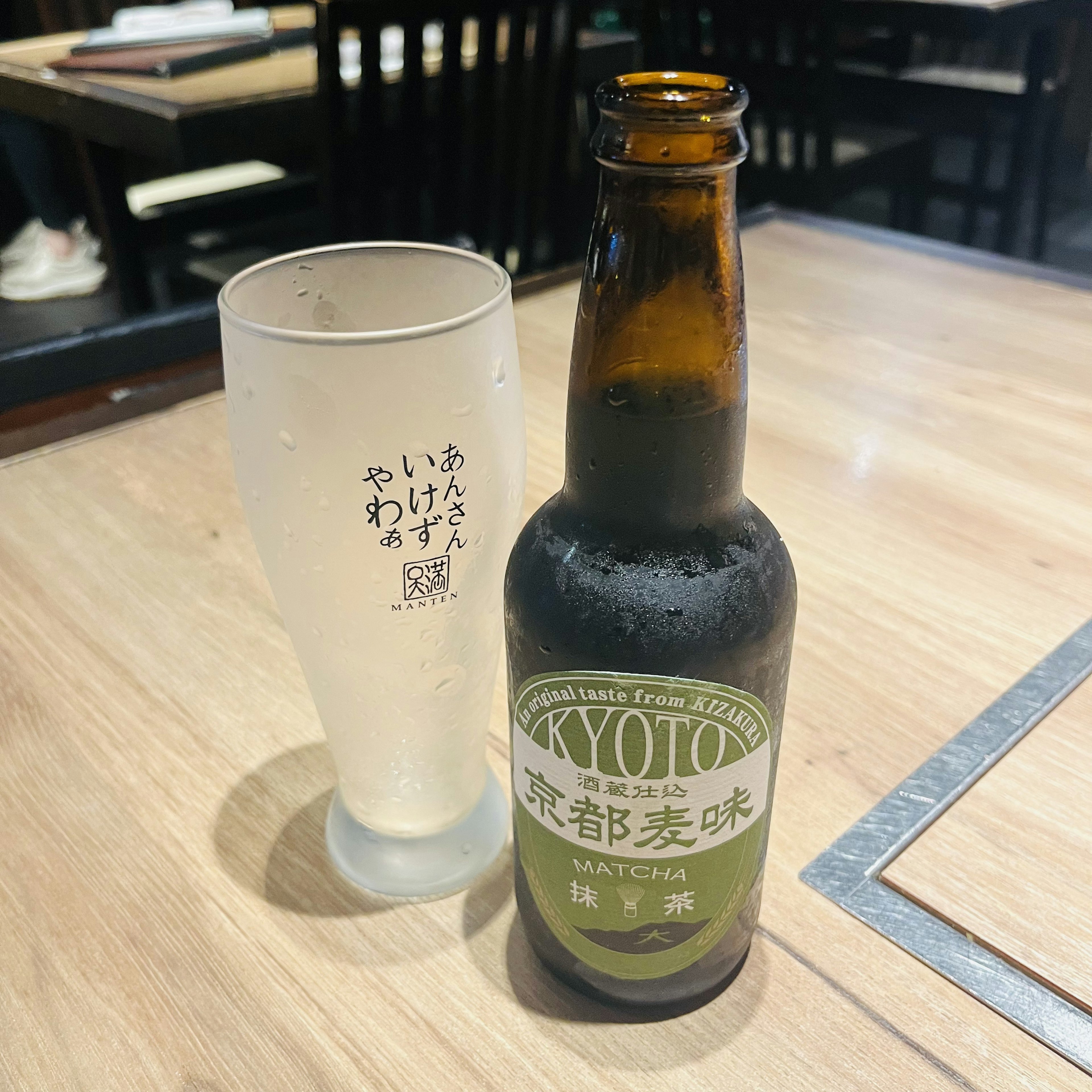 Una botella de cerveza de Kyoto junto a un vaso transparente sobre una mesa de madera