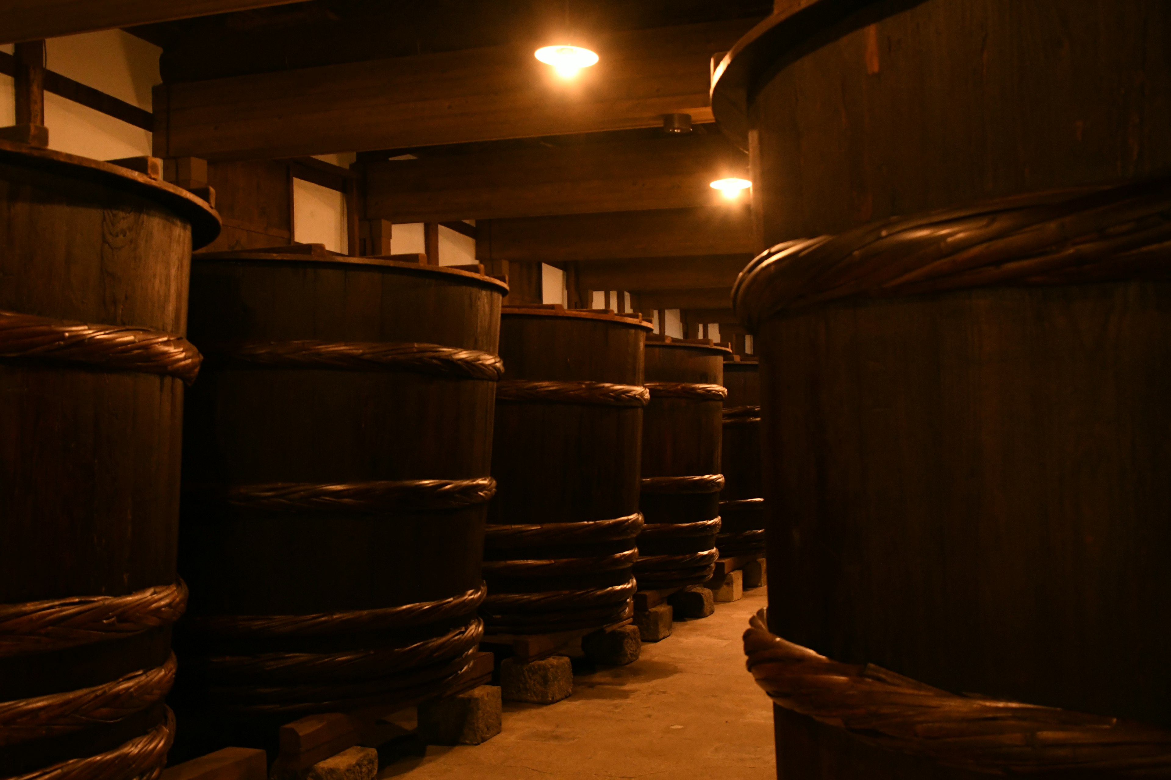 Interior of a sake brewery featuring large wooden barrels in a dimly lit space