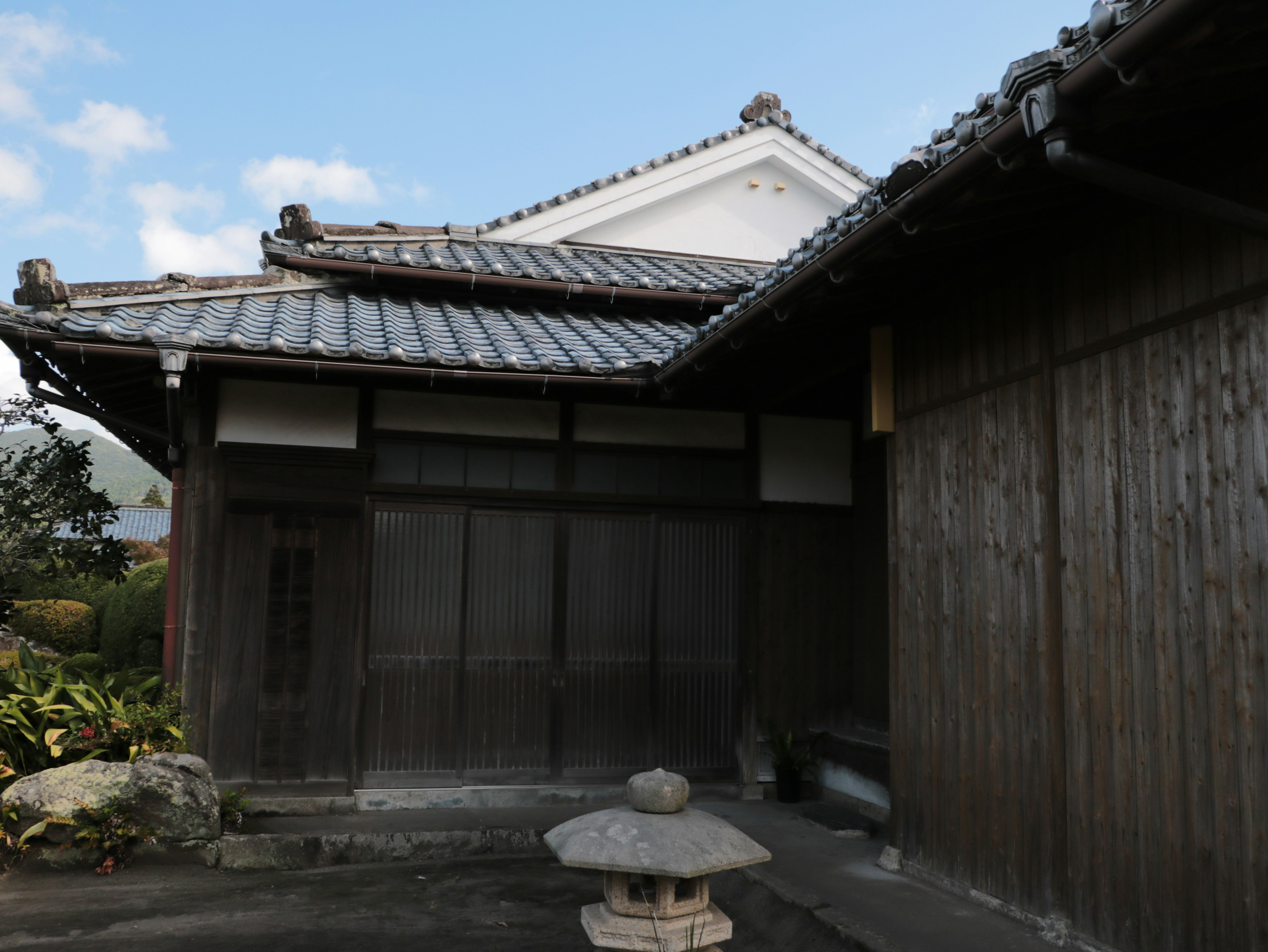 Traditional Japanese house exterior with wooden walls and tiled roof featuring a stone lantern in the garden