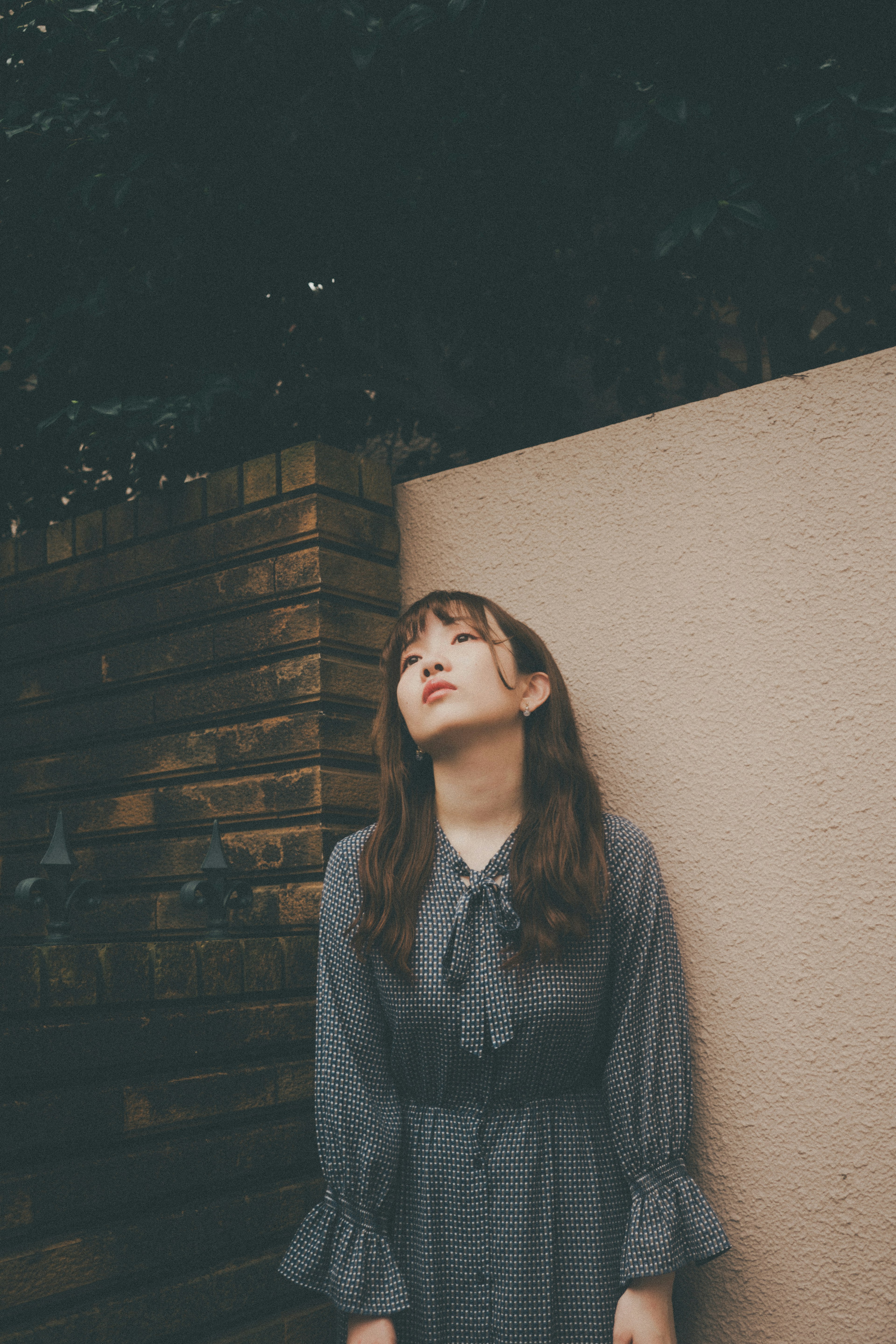 A woman in a gray dress leaning against a wall and looking up at the sky