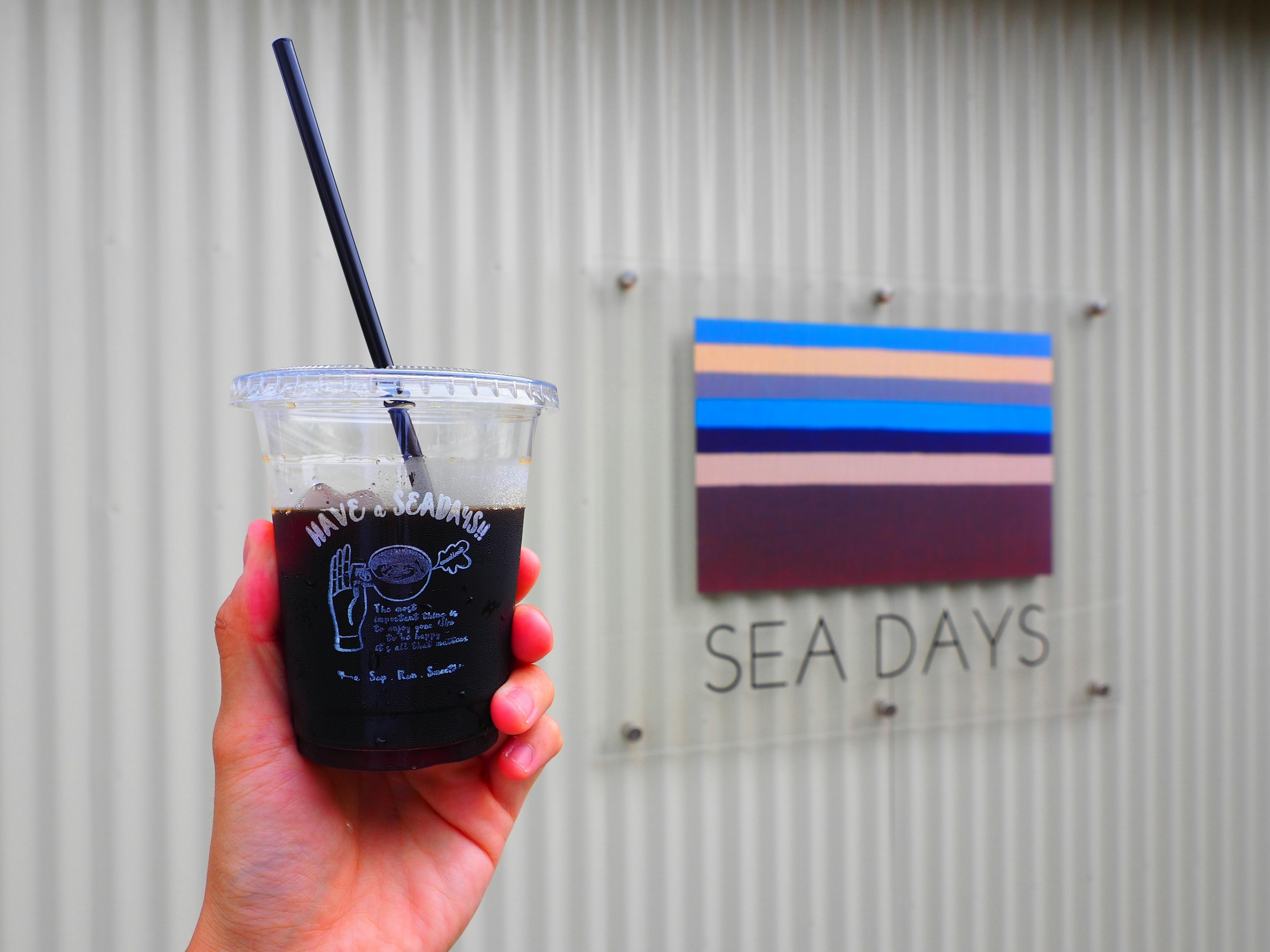 มือที่ถือแก้วใสที่มีเครื่องดื่มอยู่ข้างป้ายที่เขียนว่า SEA DAYS