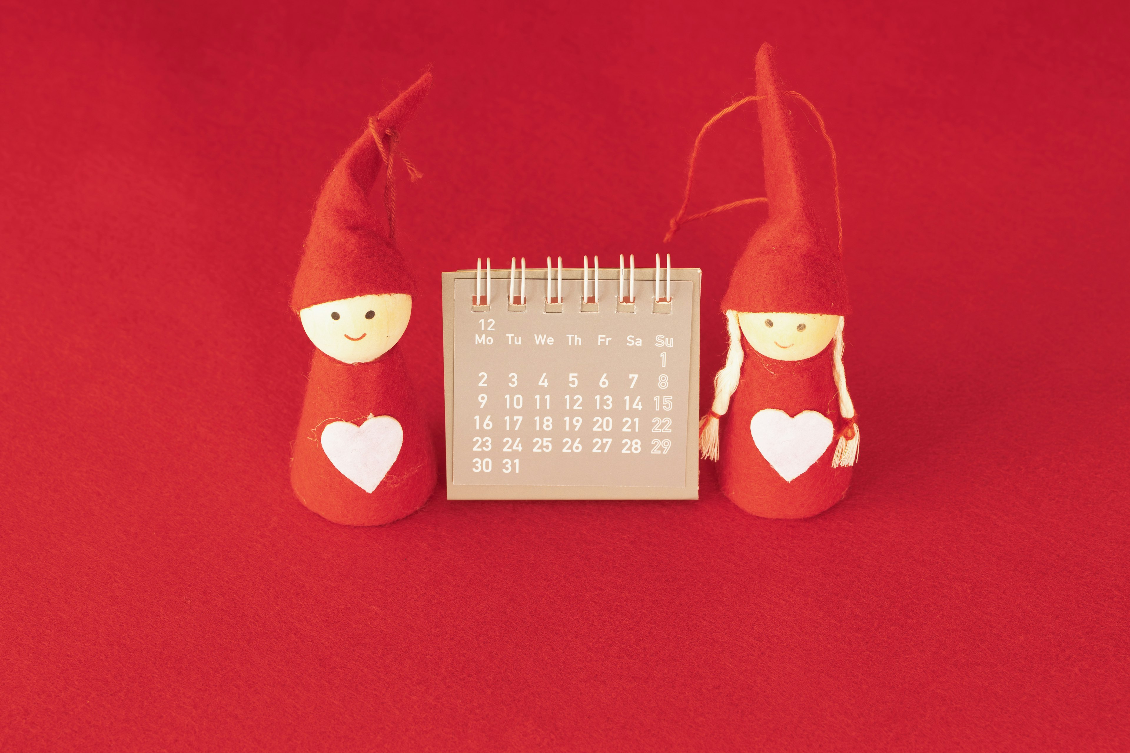 Two red-hatted dolls with hearts beside a calendar on a red background