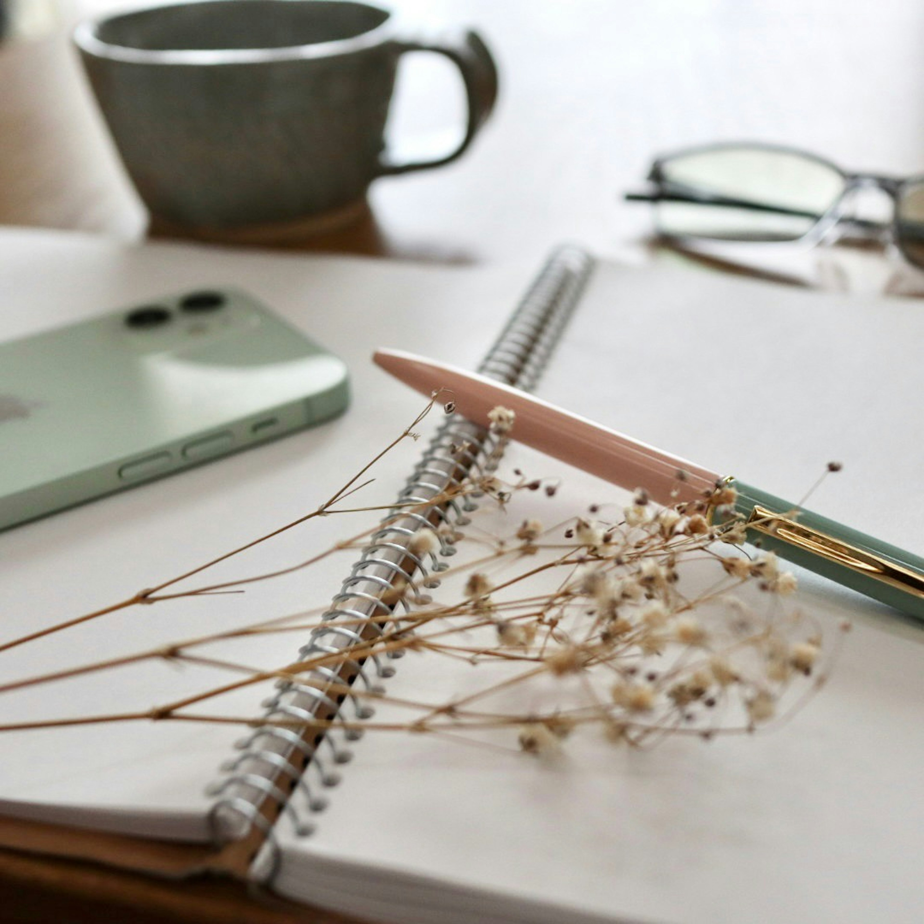 Eine ruhige Schreibtischszene mit einer grünen Tasse, Smartphone, Spiralnotizbuch, Stift und Trockenblumen