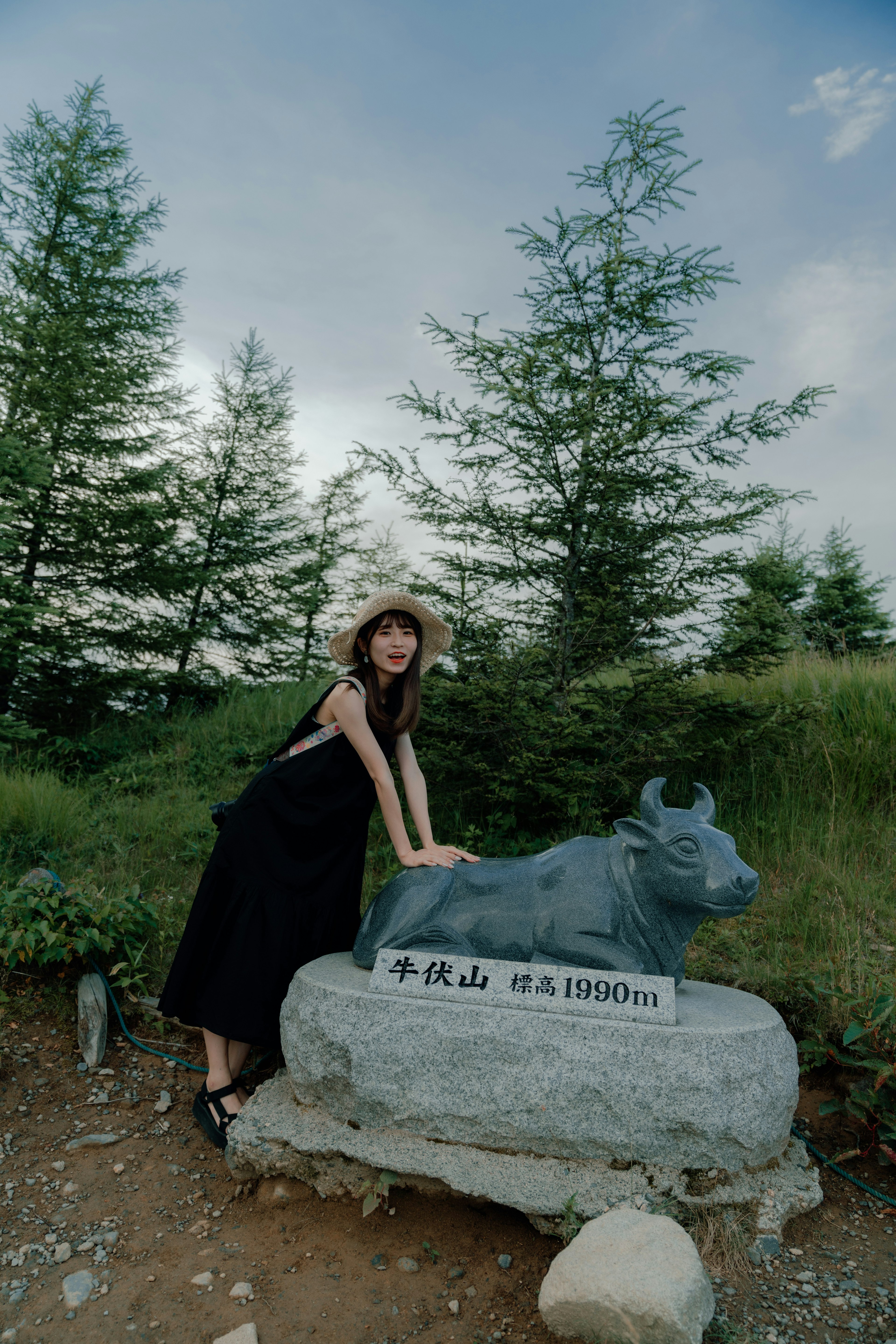 A woman in a black dress posing next to a stone cow statue in a grassy area