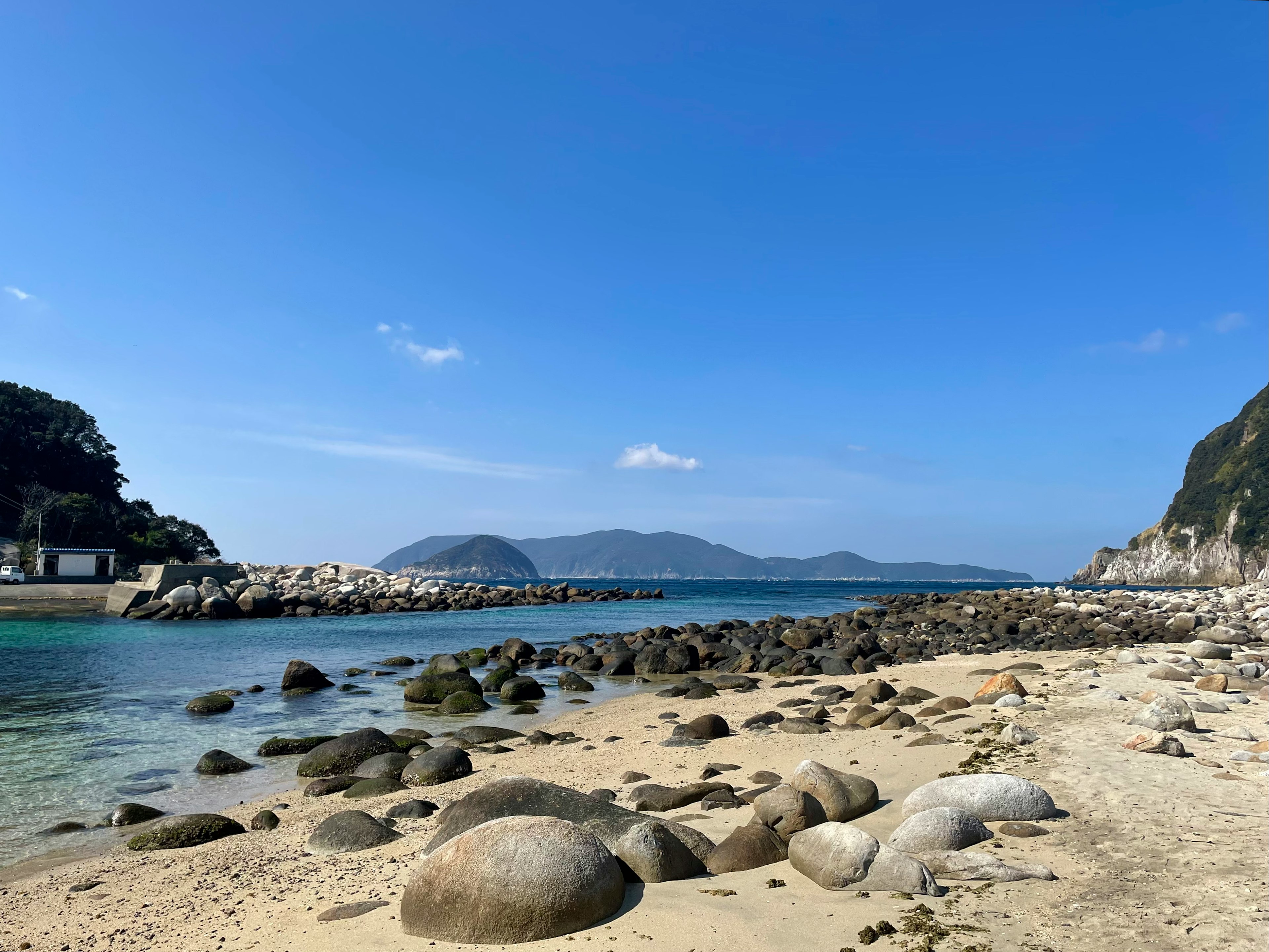 푸른 하늘과 잔잔한 바다가 펼쳐진 해변 경관 해변에 흩어진 큰 돌들 배경에 보이는 산들