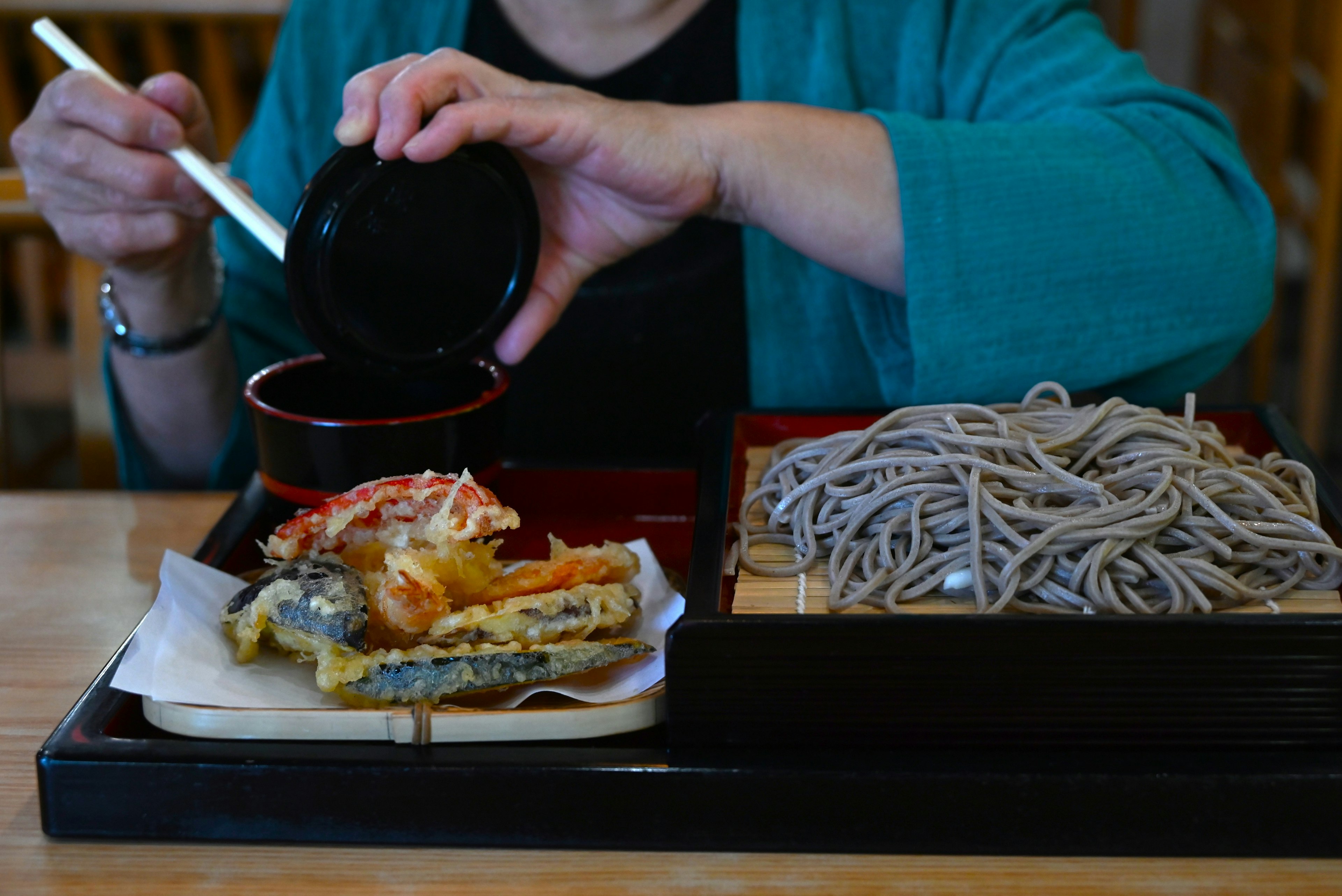 A hand pouring sauce over tempura and soba noodles