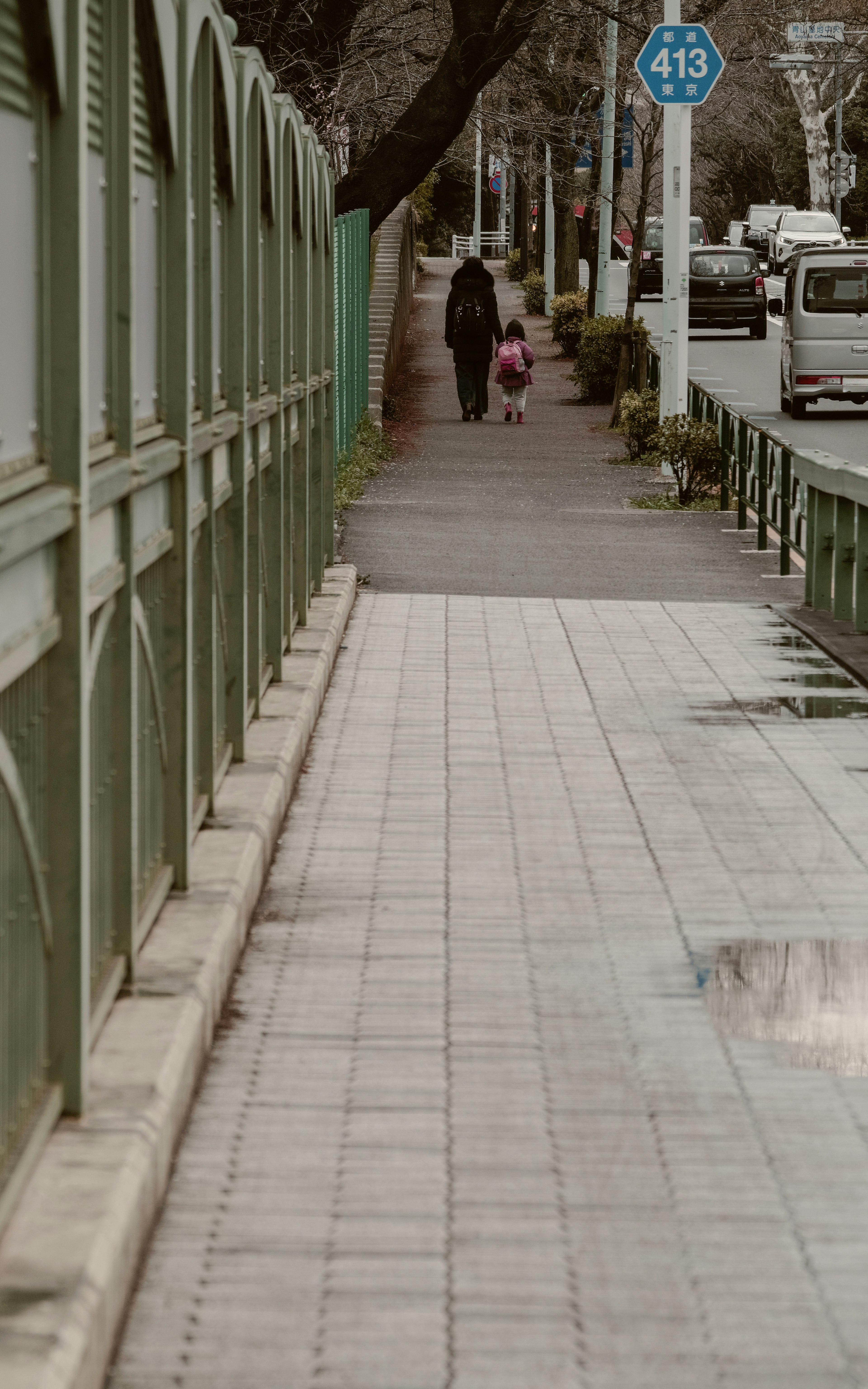 Seorang orang tua dan anak berjalan di trotoar dengan pagar hijau dan lampu jalan