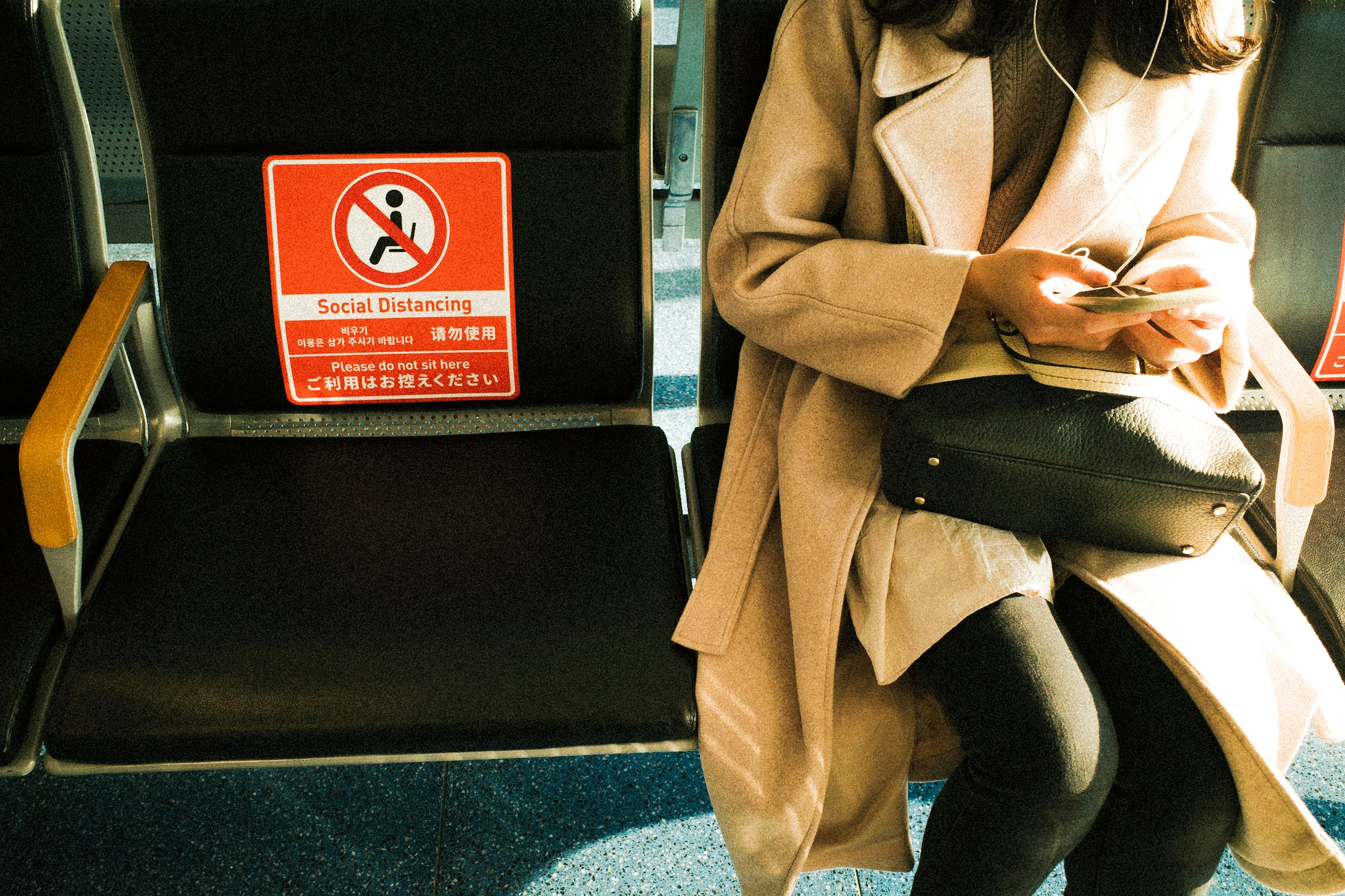 Close-up of a woman sitting with a phone in hand wearing a beige coat next to a warning sign