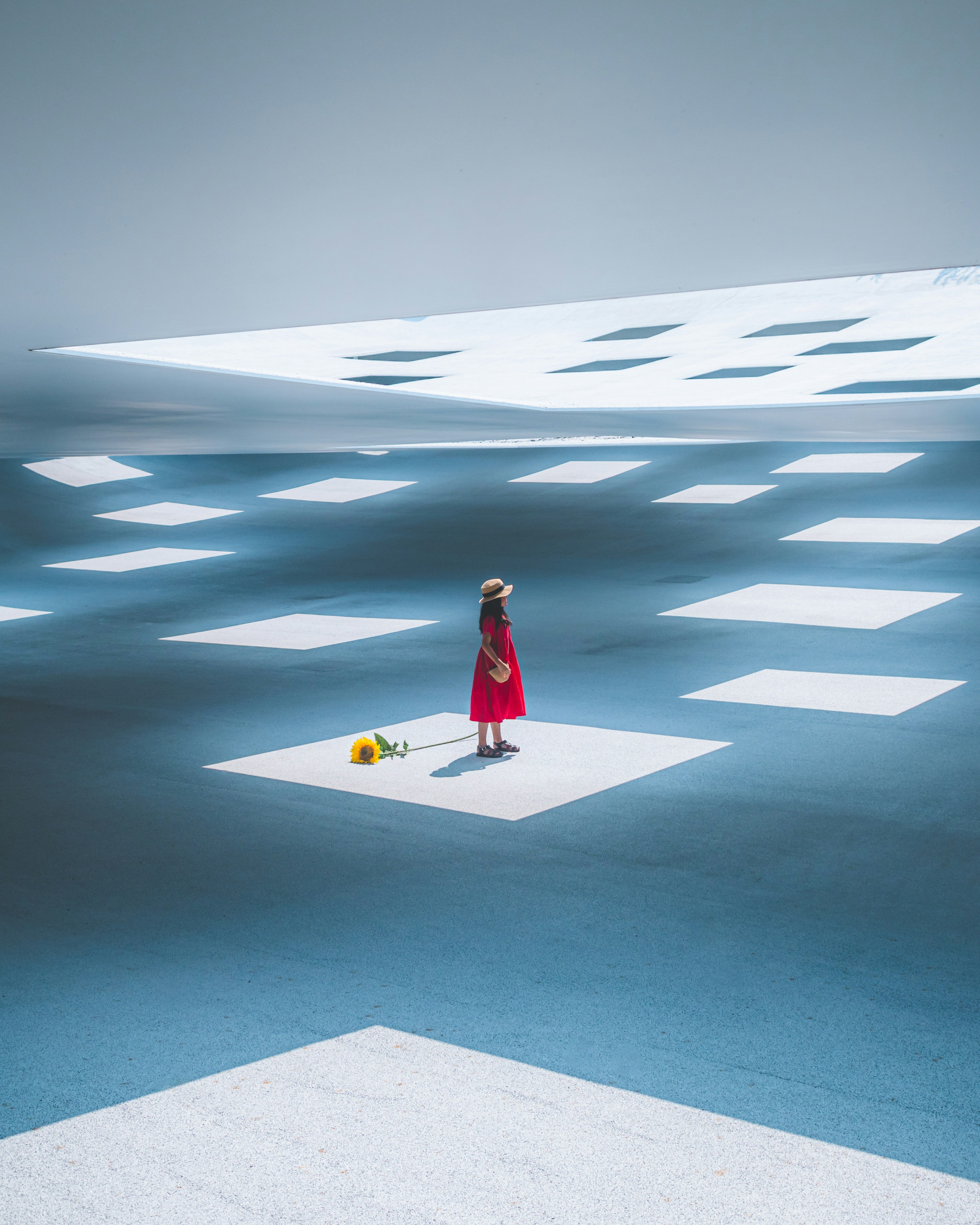 Une fille en robe rouge se tient sur un sol bleu entourée de grands motifs carrés blancs