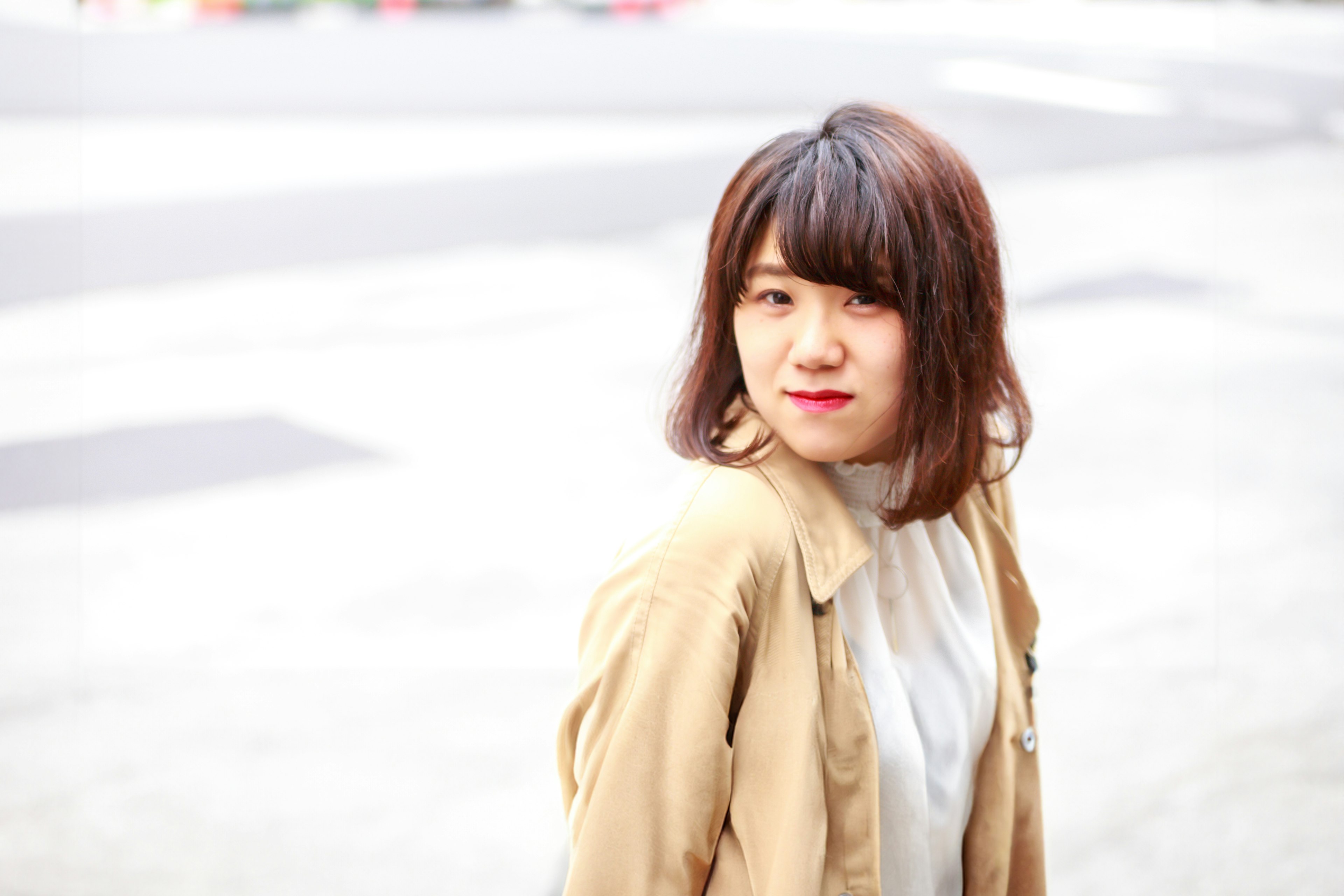 A woman smiling and looking back wearing a white blouse and beige jacket with a blurred urban background