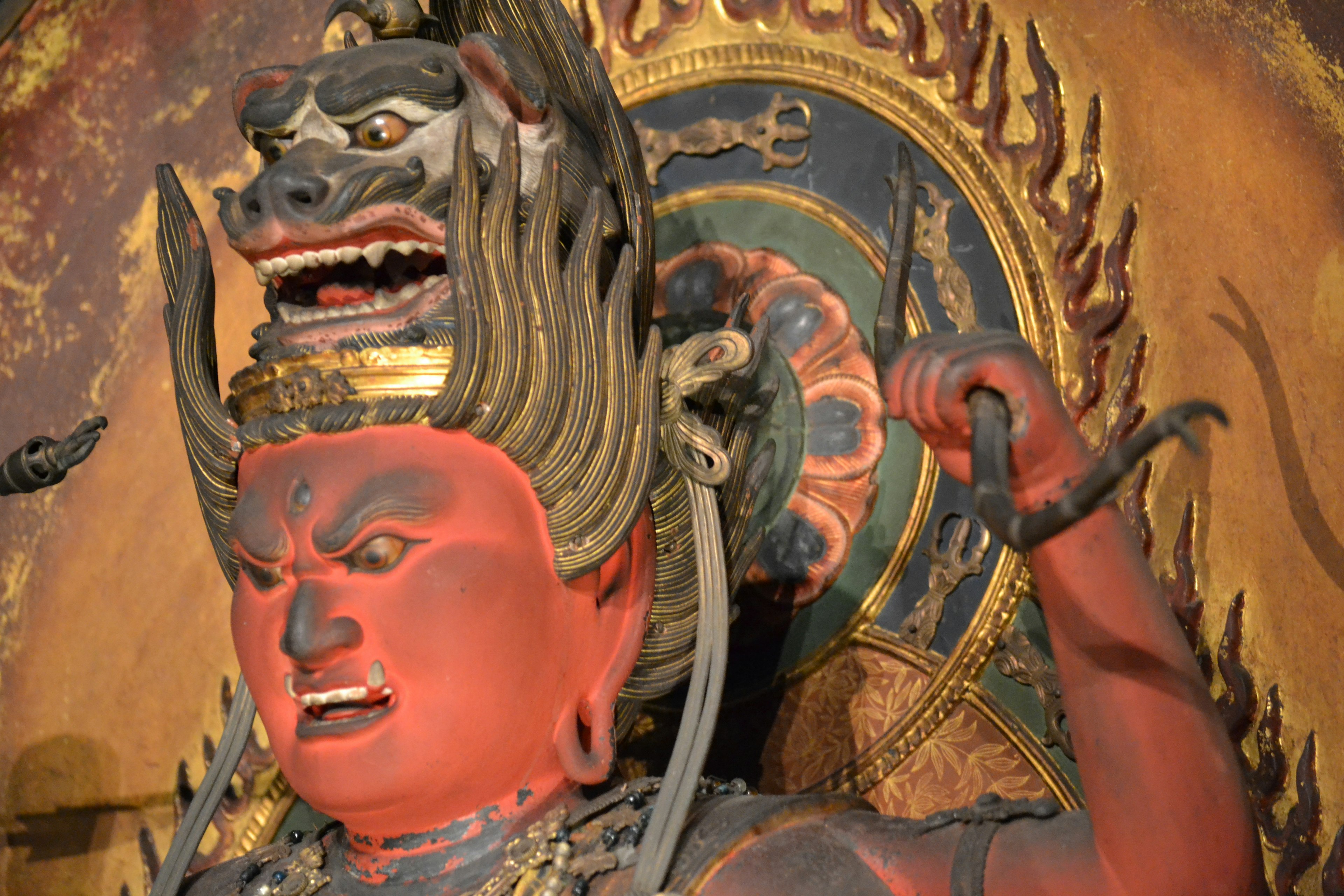 A red-faced deity statue displaying an angry expression with a decorative golden symbol in the background