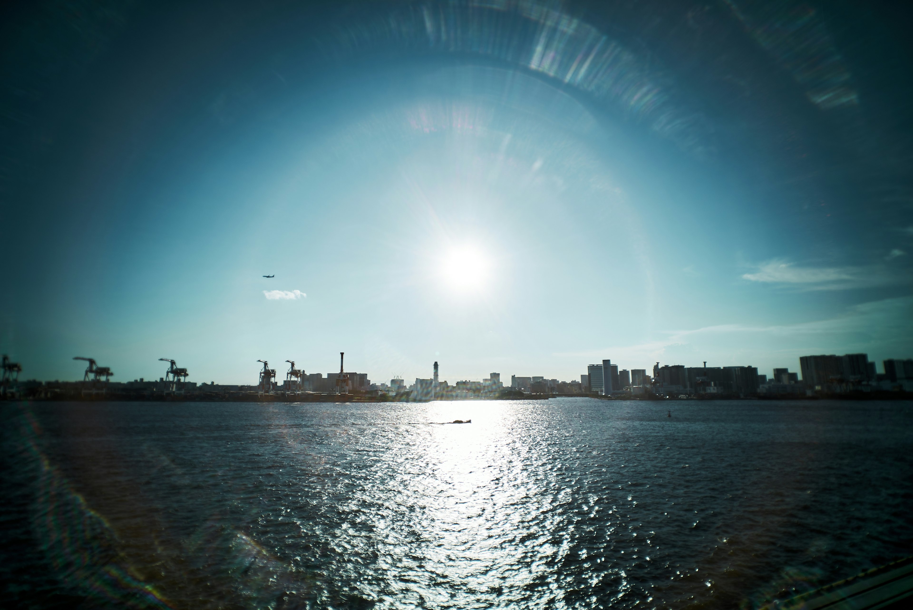 Landscape featuring a bright sun reflecting on water with cranes in the harbor