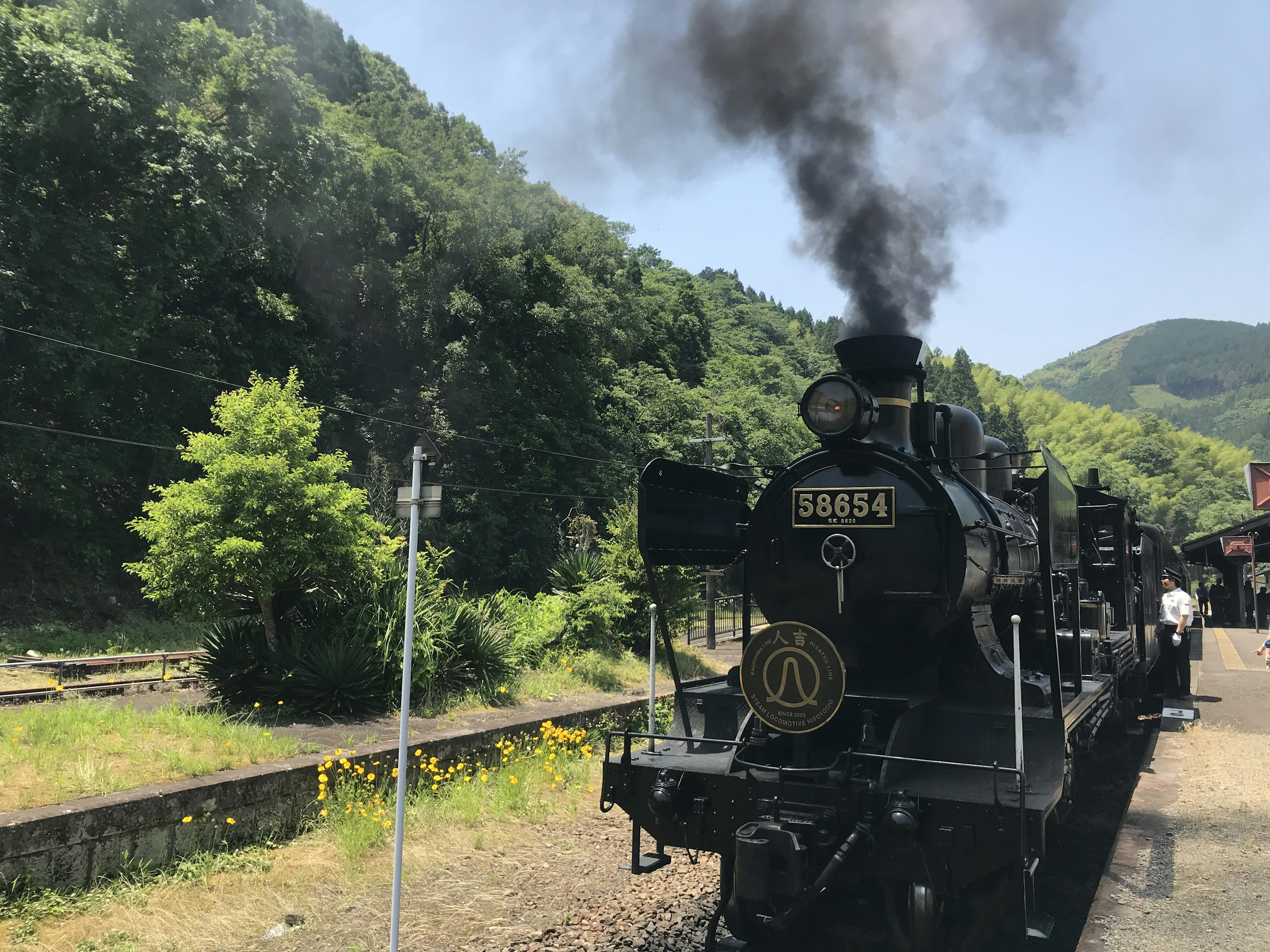 Schwarze Dampflokomotive, die Rauch ausstößt, mit grünen Bergen und Bäumen im Hintergrund