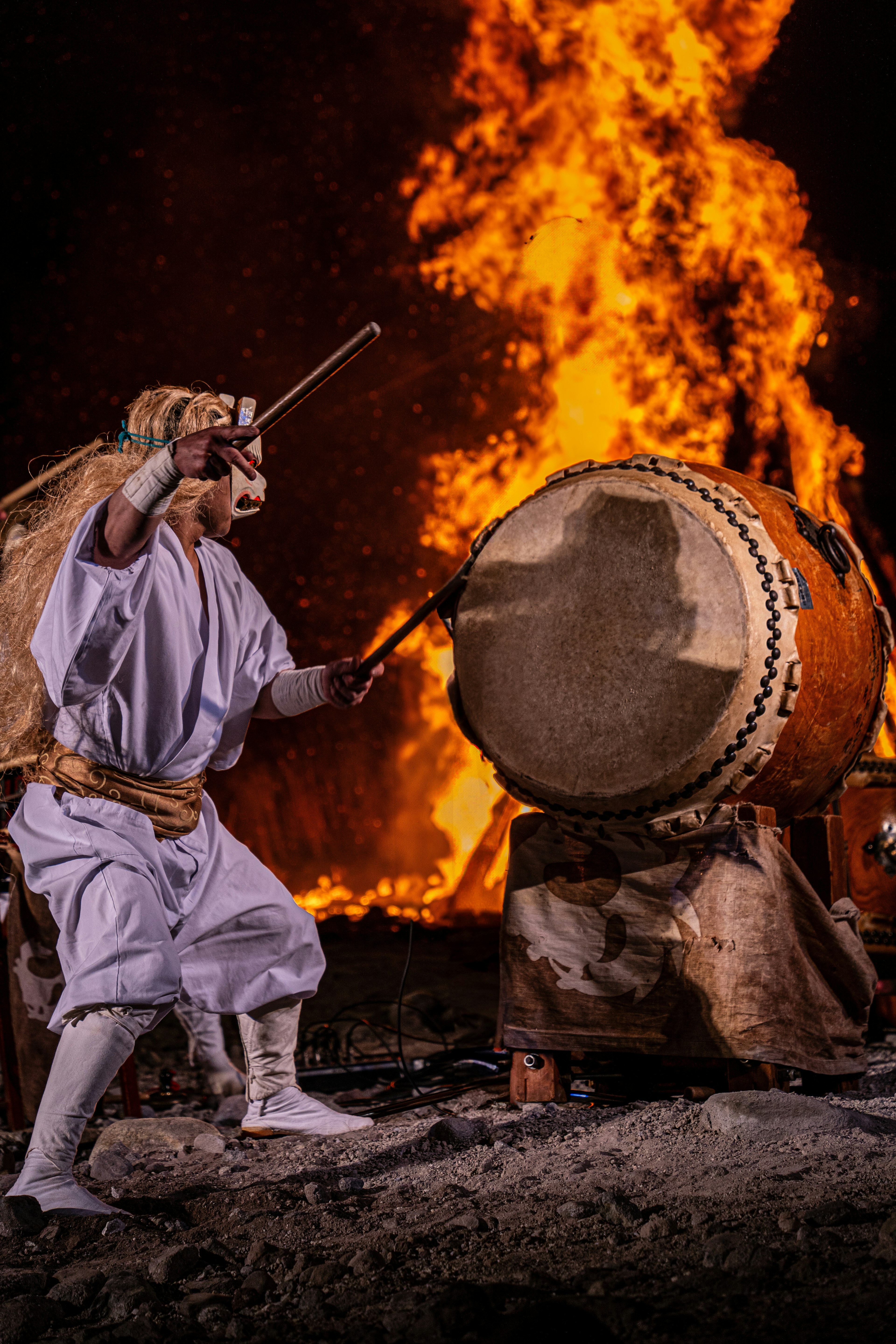 Man in traditional attire playing a drum with fire in the background