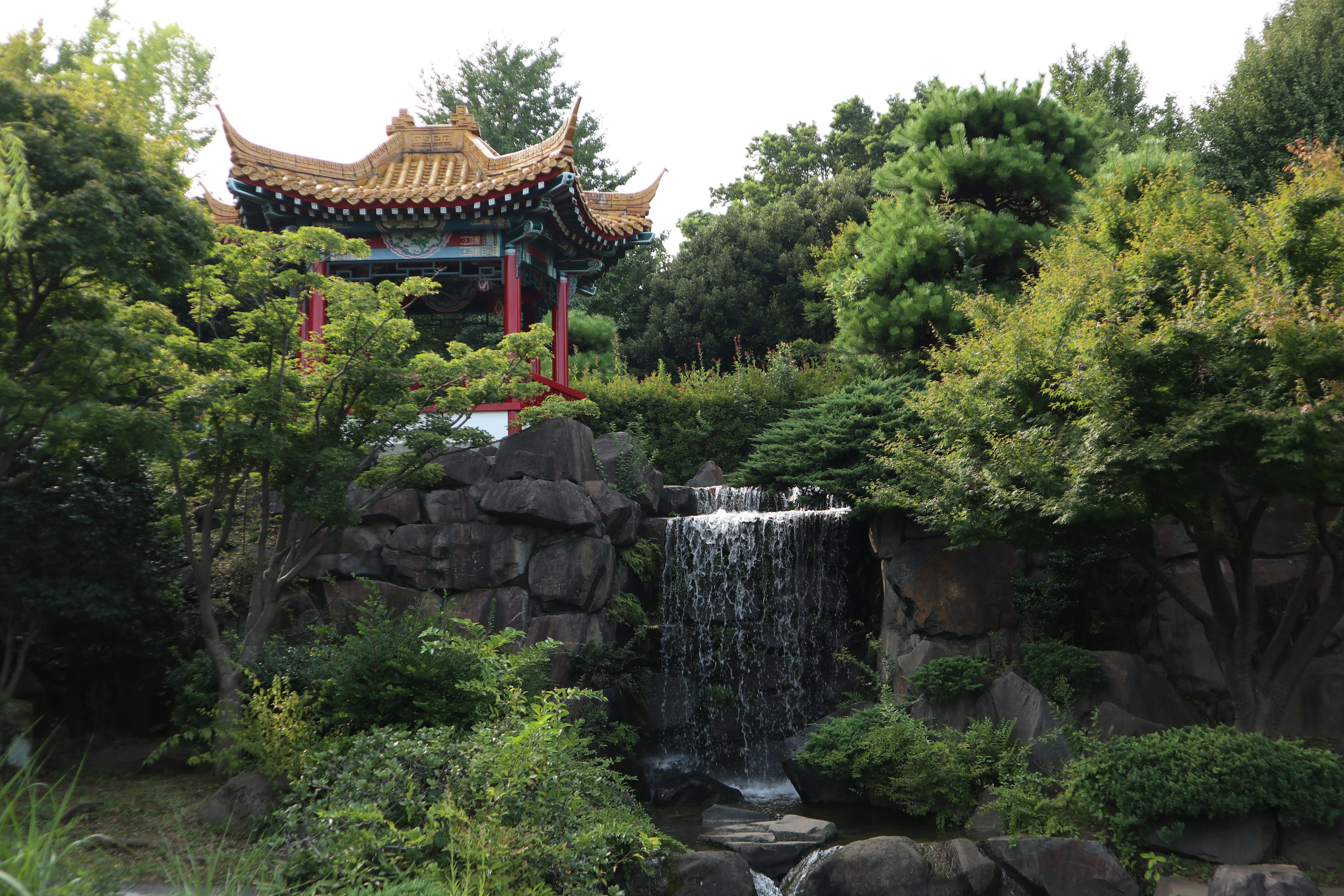 Chinese-style pavilion surrounded by lush greenery and a small waterfall in a Japanese garden