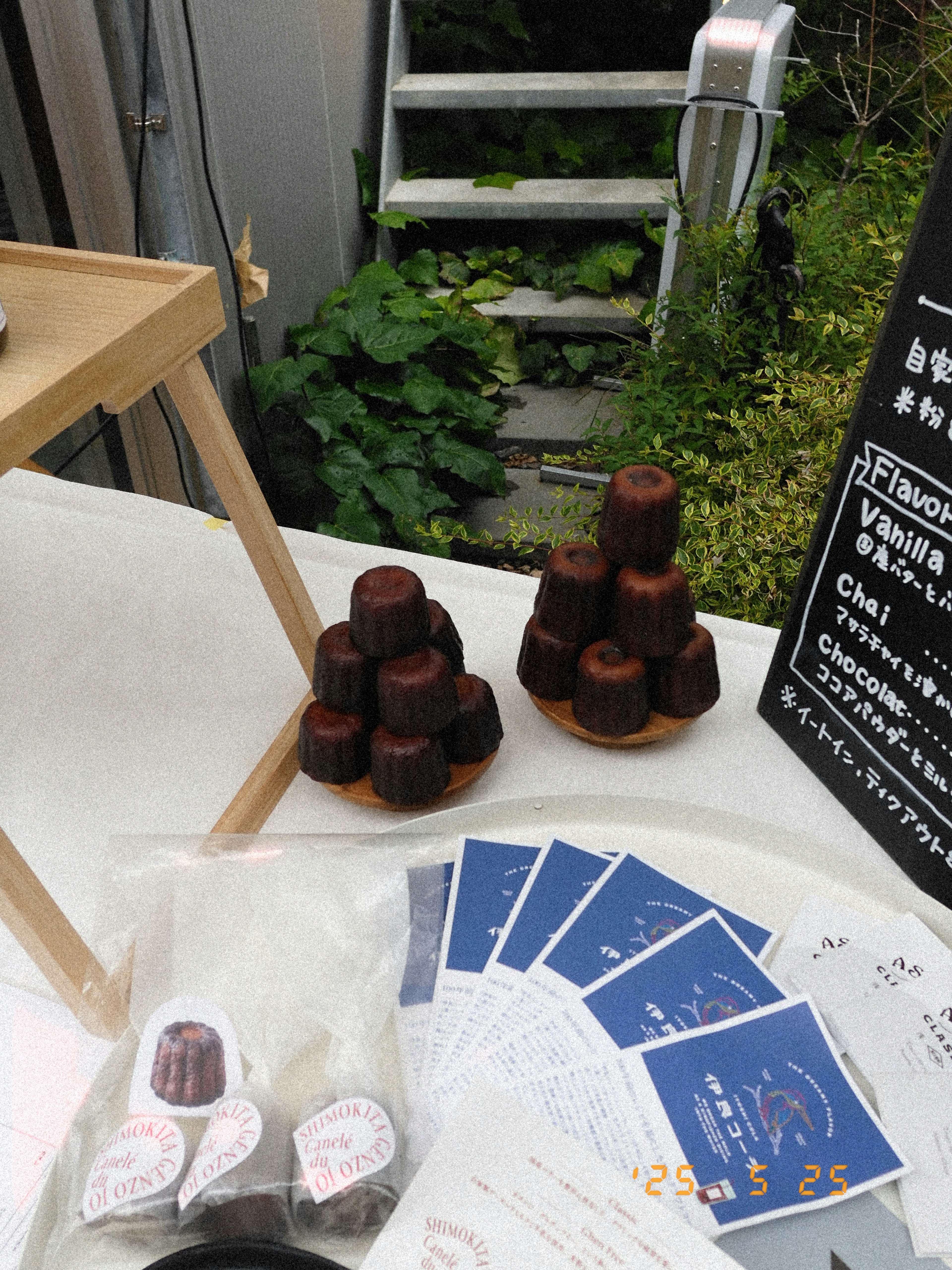 Exhibición de canelés en una mesa con tarjetas azules y vegetación al fondo