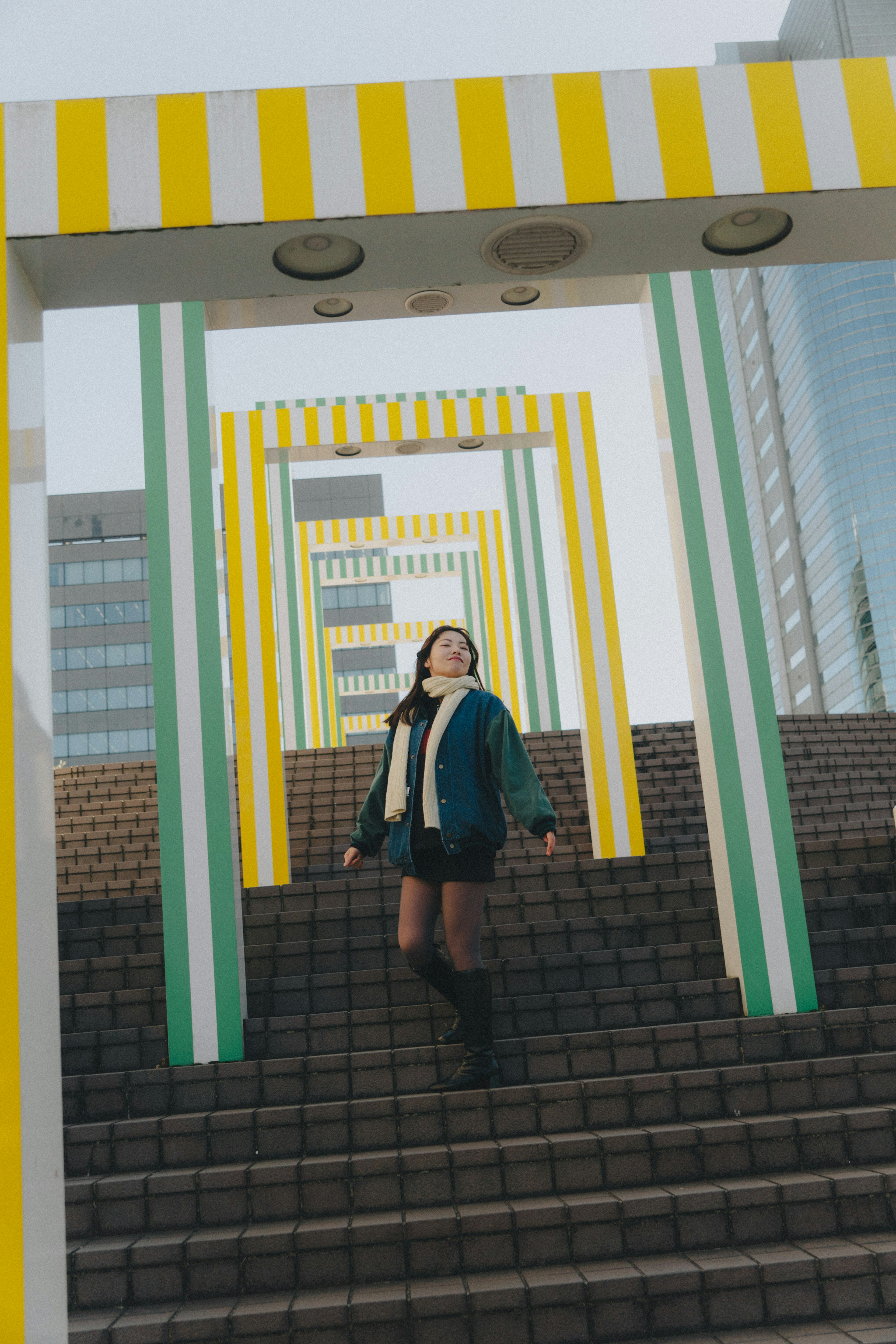 A woman in a blue jacket poses joyfully on stairs with colorful arches in the background