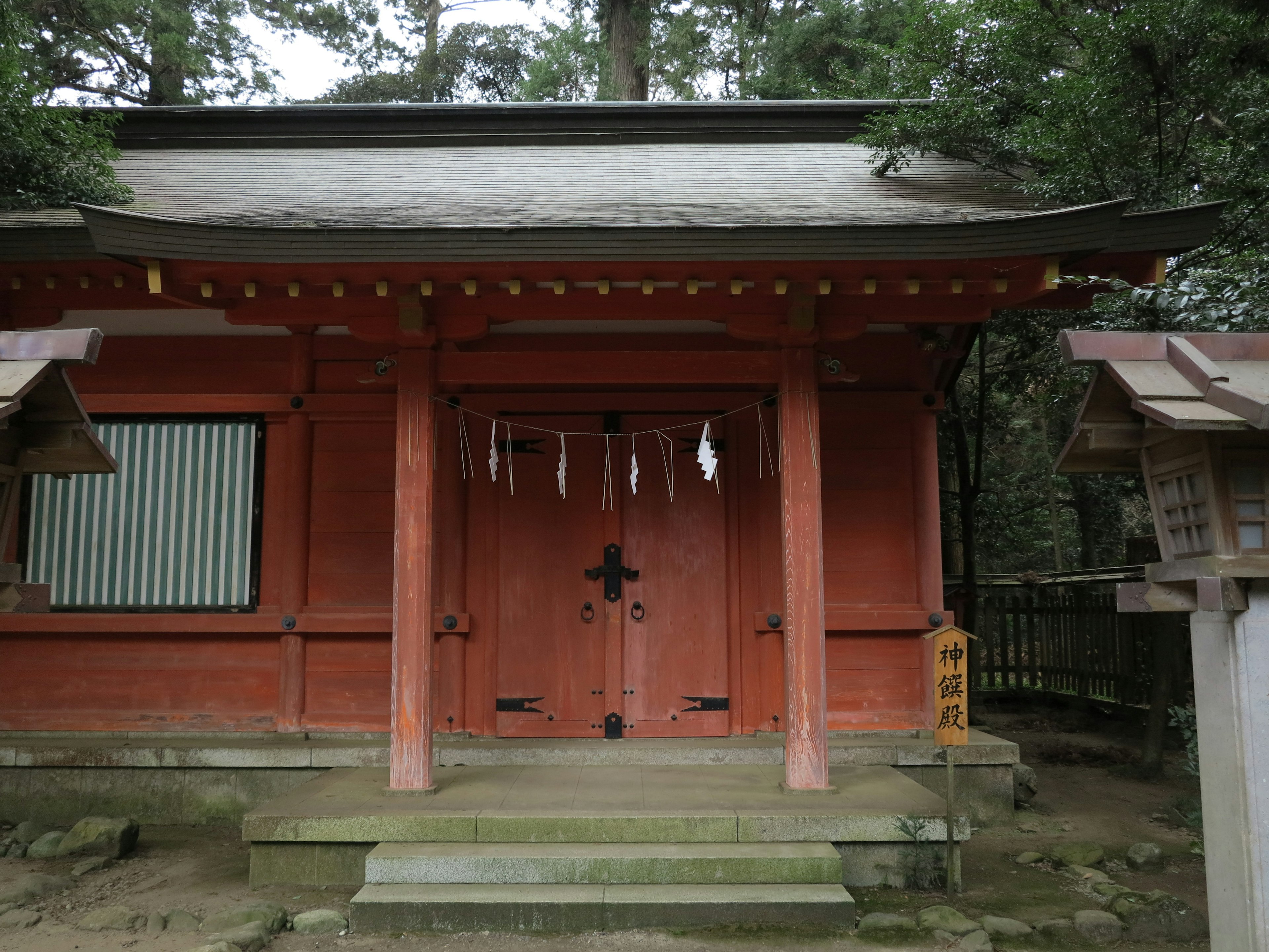 A red shrine building nestled in the forest featuring a large door and decorative elements