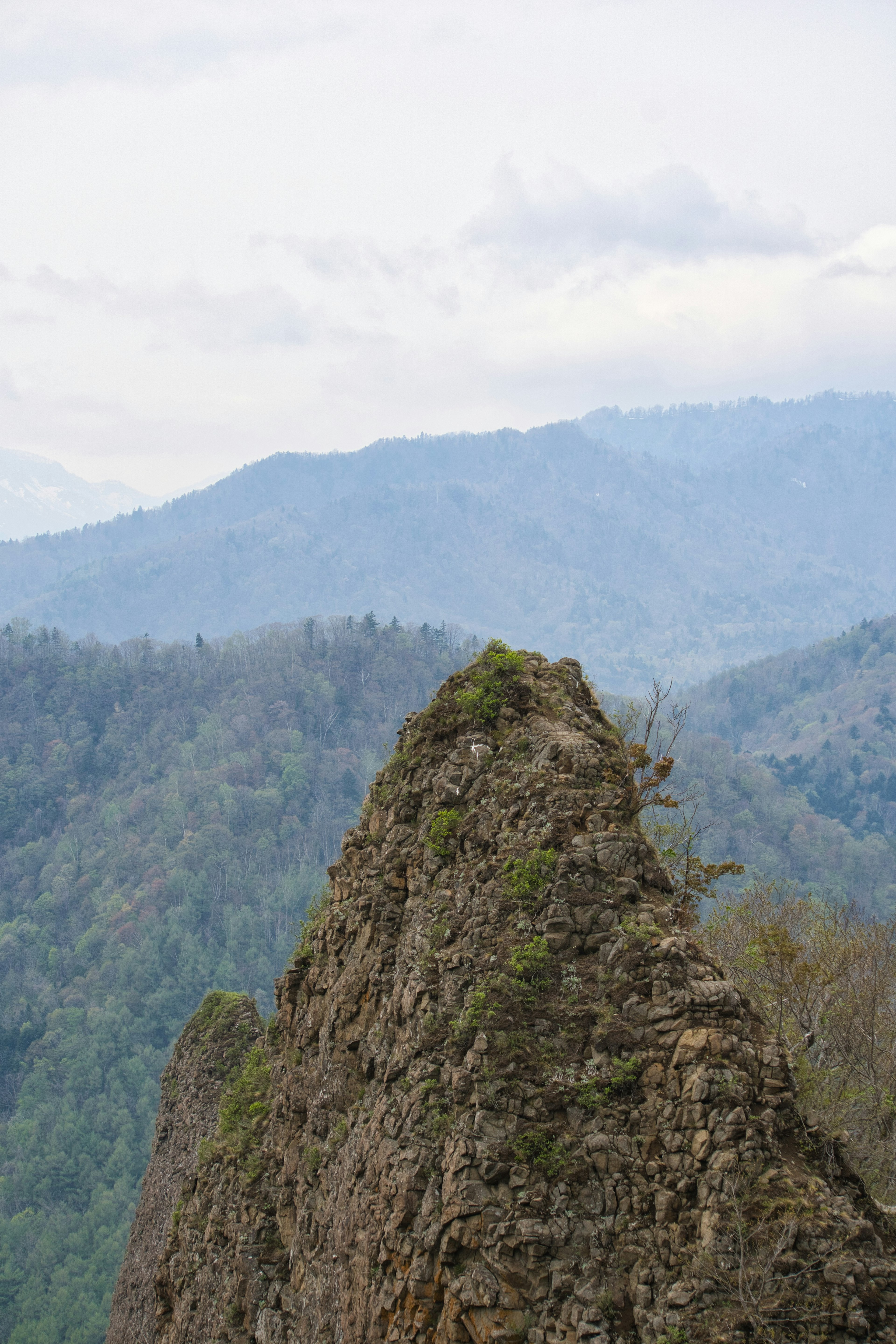 Puncak gunung yang tajam dikelilingi pohon hijau dan gunung jauh