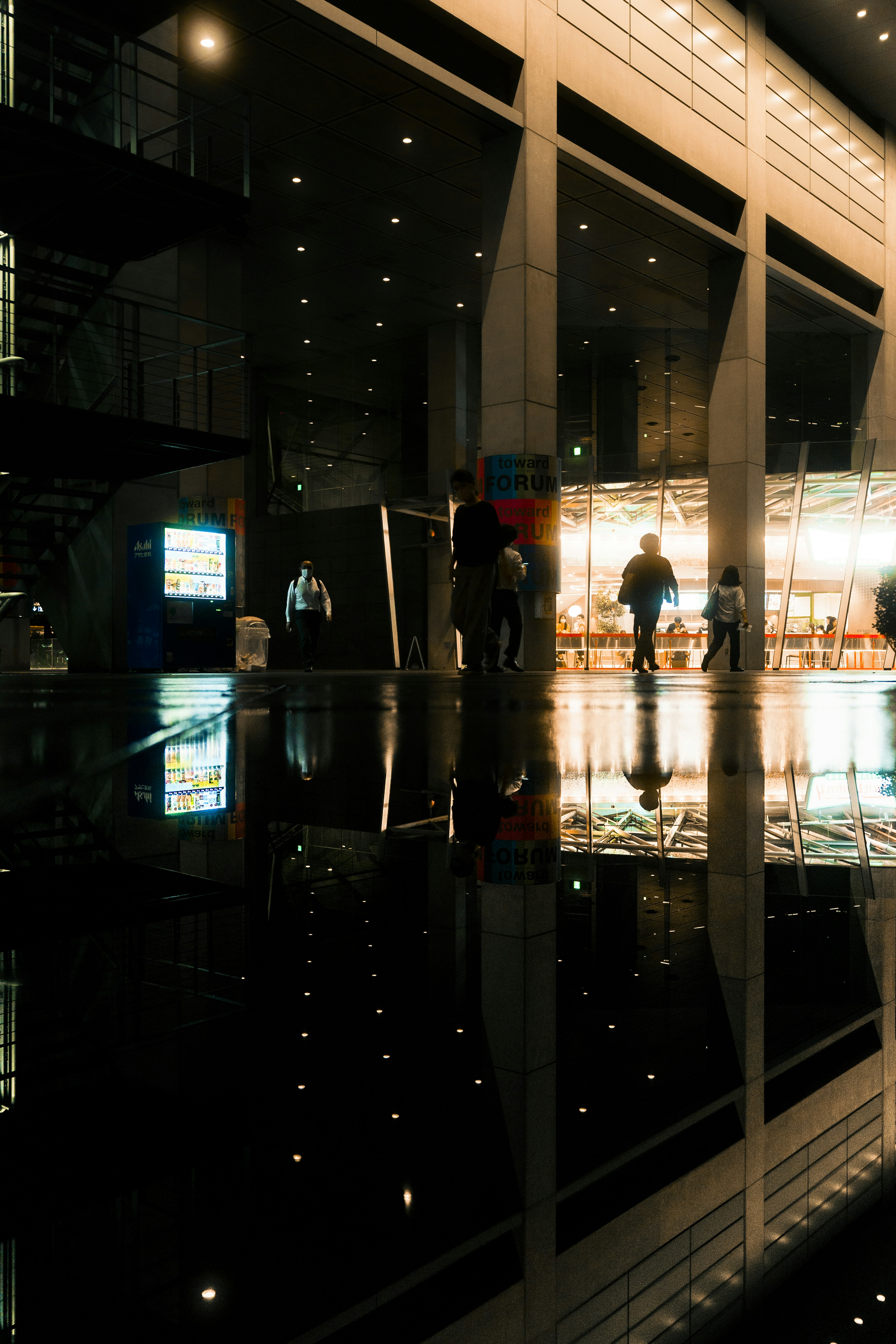 現代的な建物の外観と人々の影が水たまりに映るシーン