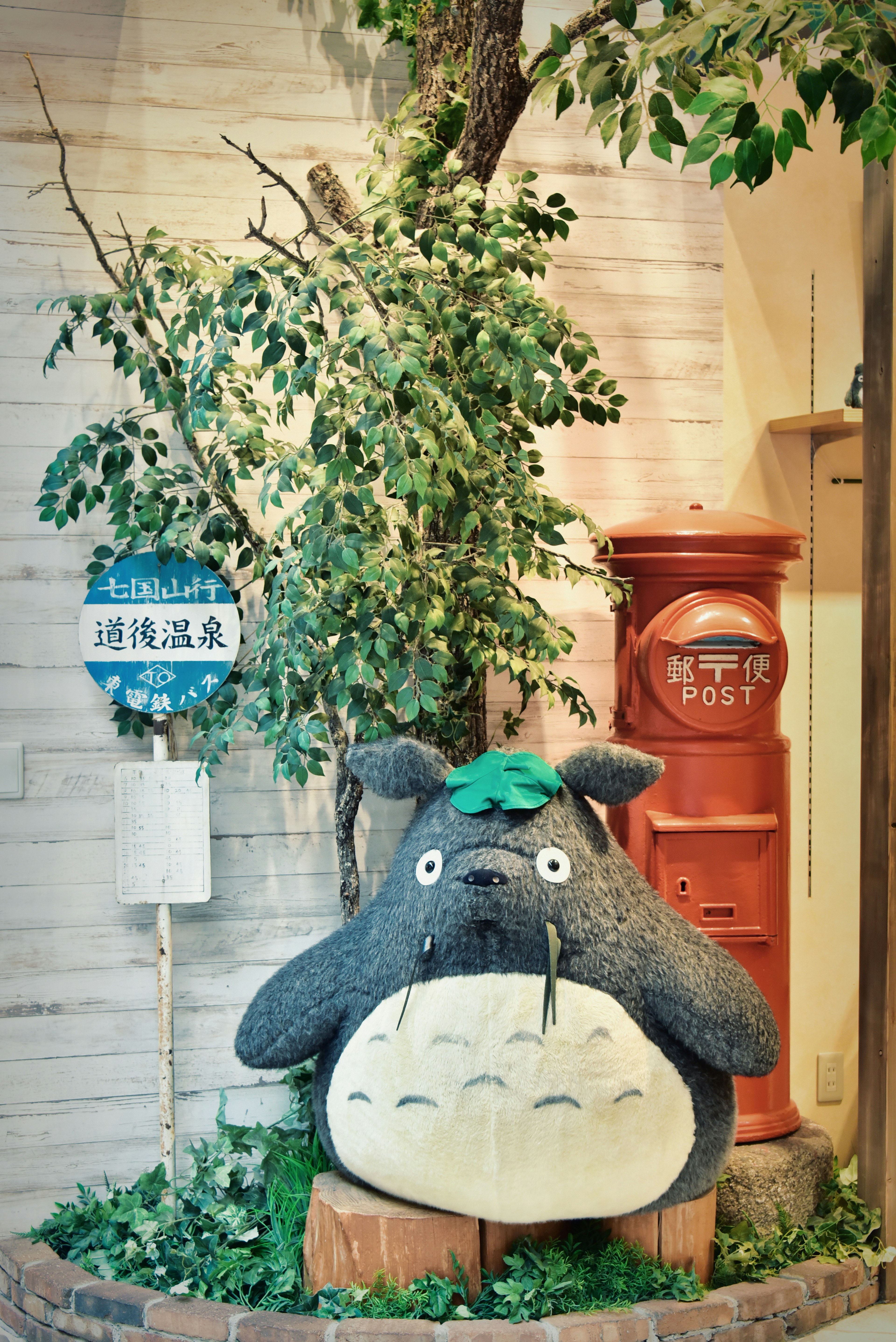 Ein großer Totoro-Plüschspielzeug umgeben von Grün mit einem roten Briefkasten im Hintergrund