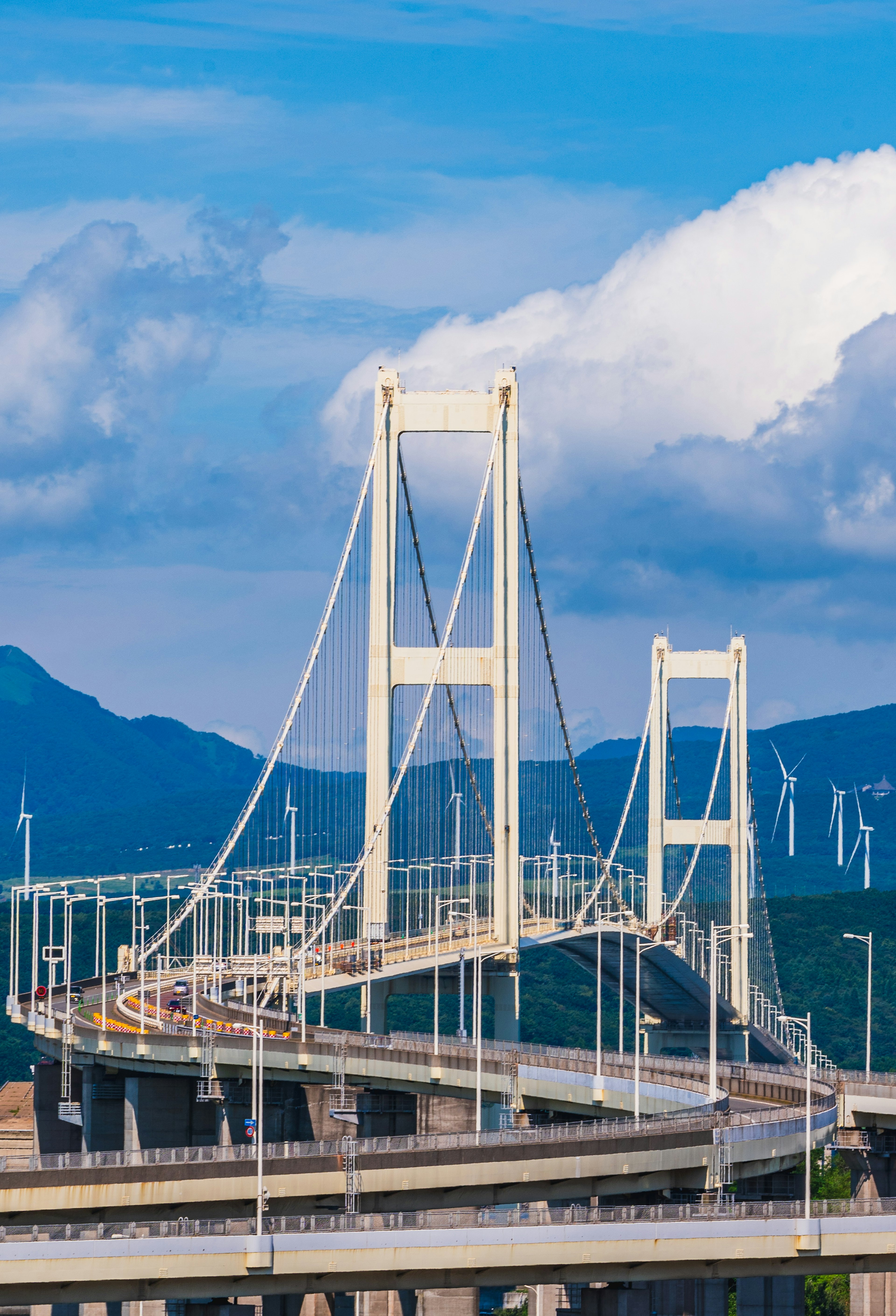 Eine weiße Hängebrücke vor einem blauen Himmel mit Wolken Straßen und Berge im Hintergrund