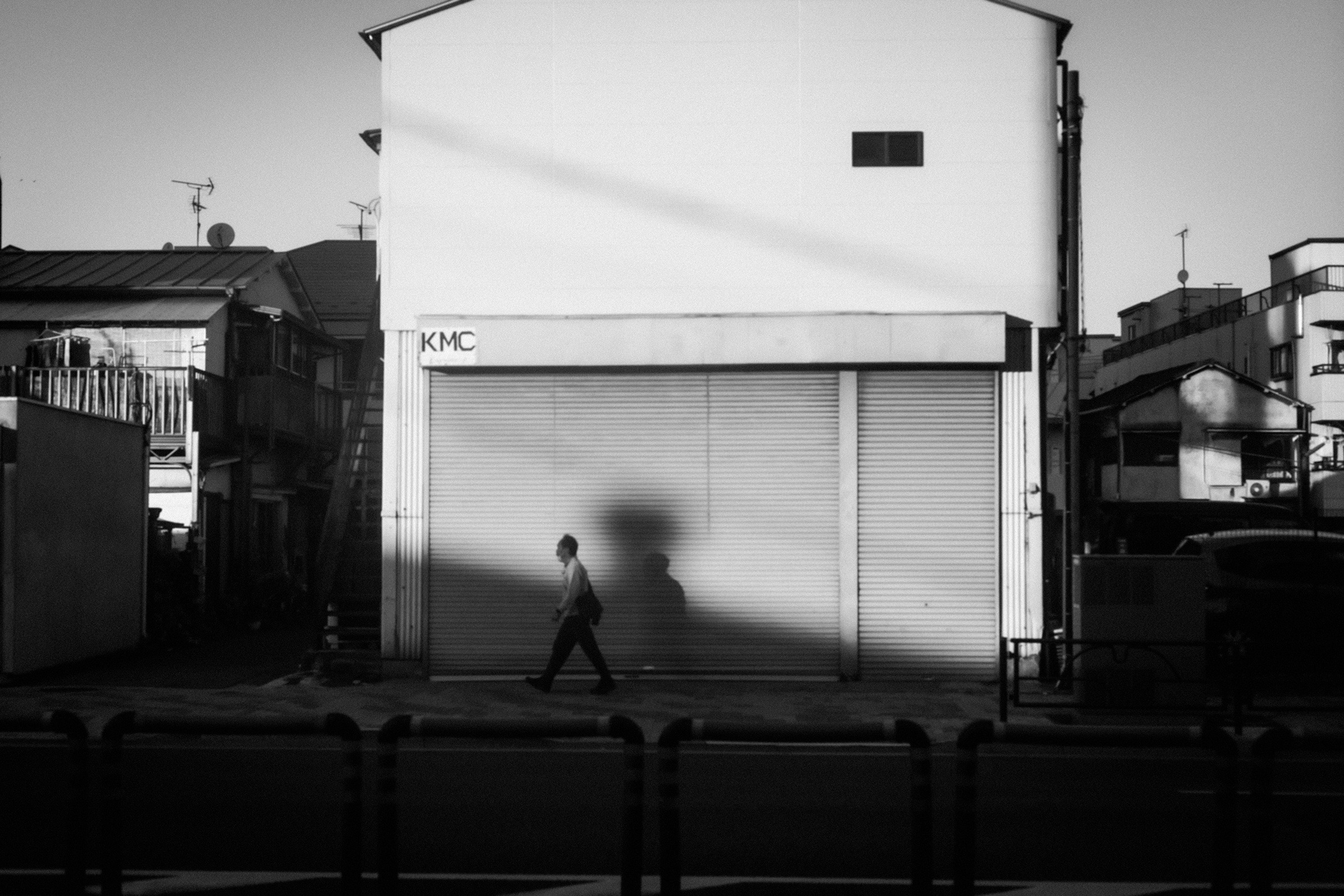 Ein Schwarzweißfoto mit einer Person, die vor einem weißen Gebäude mit einem auffälligen Schatten geht