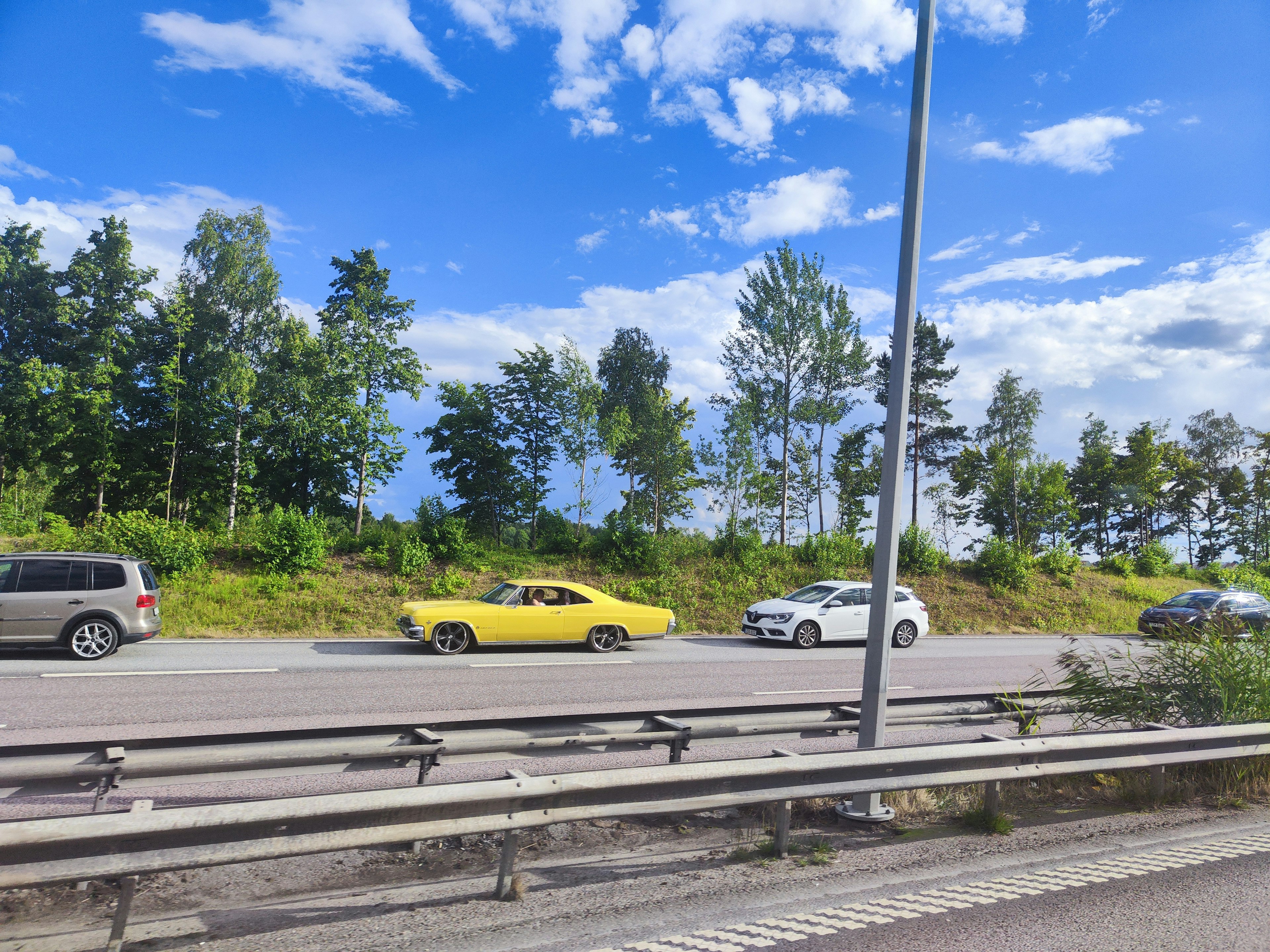 Autobahnszene mit einem gelben Auto und einem weißen Auto umgeben von grünen Bäumen und blauem Himmel