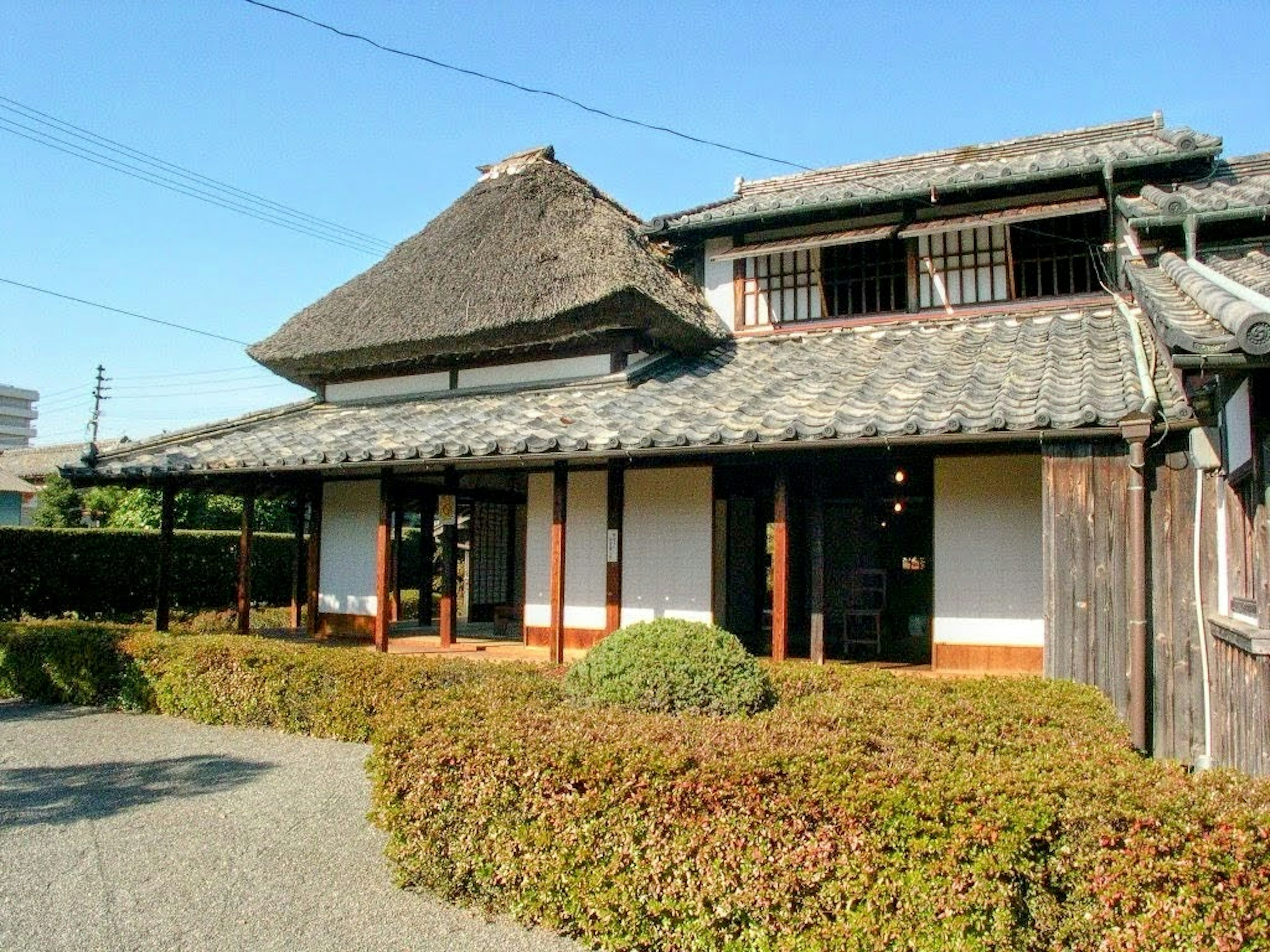 Extérieur d'une maison japonaise traditionnelle avec un toit de chaume et des murs en bois entourés de buissons verts et d'un ciel bleu