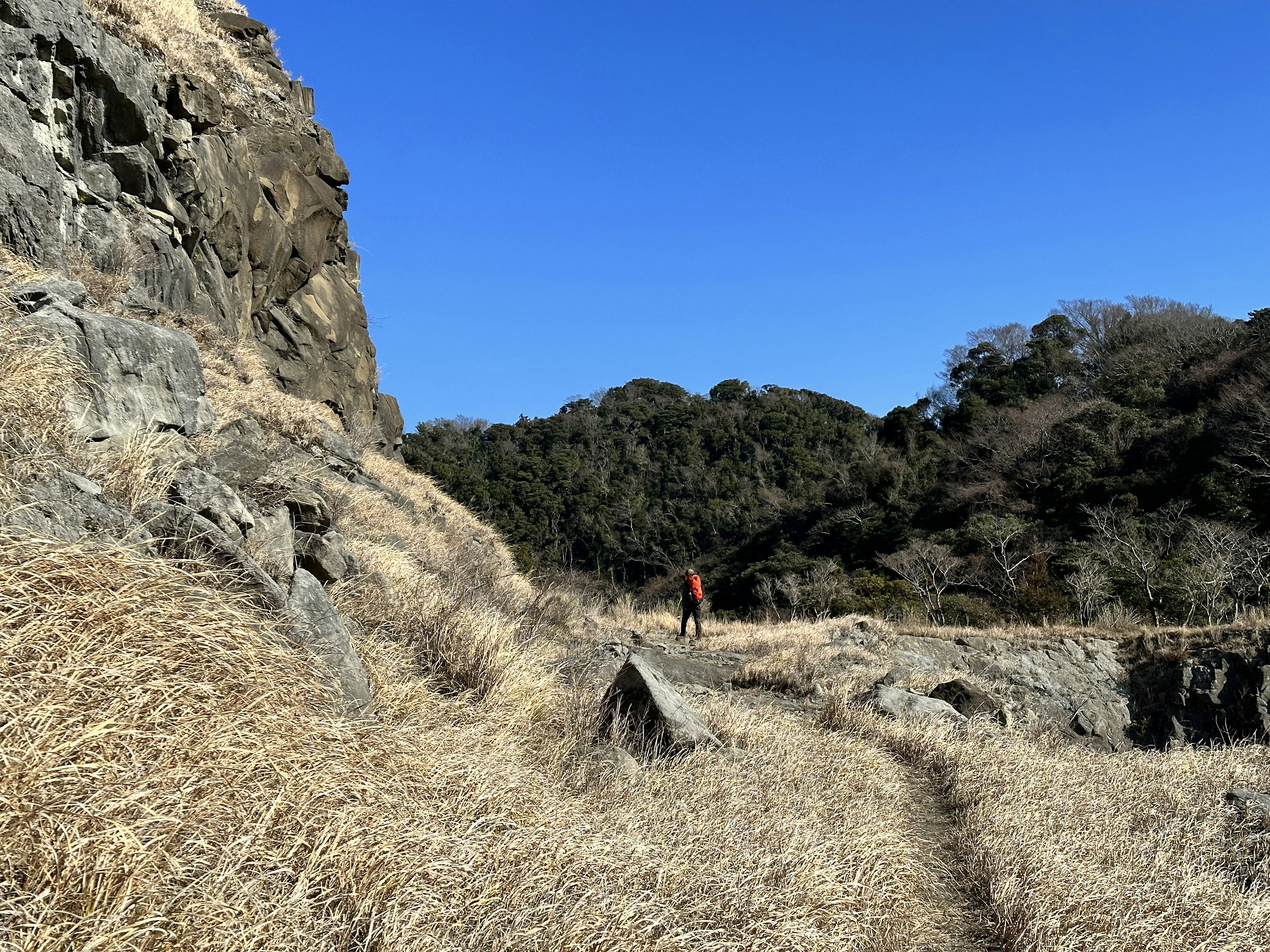 Senderista caminando en un área de hierba bajo un cielo azul con acantilados rocosos