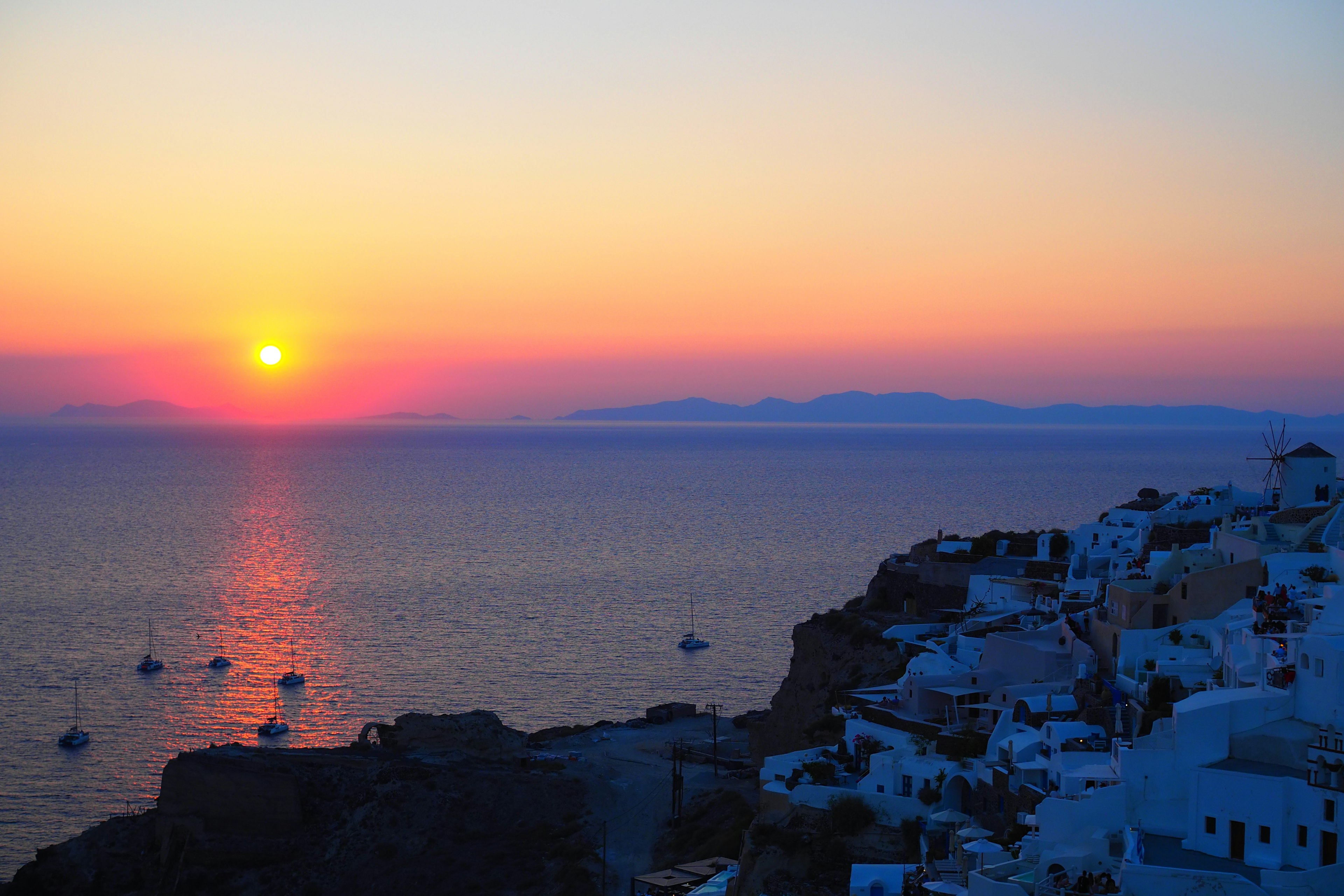 Schöner Sonnenuntergang über dem Meer in Santorin mit sichtbaren weißen Gebäuden und Booten