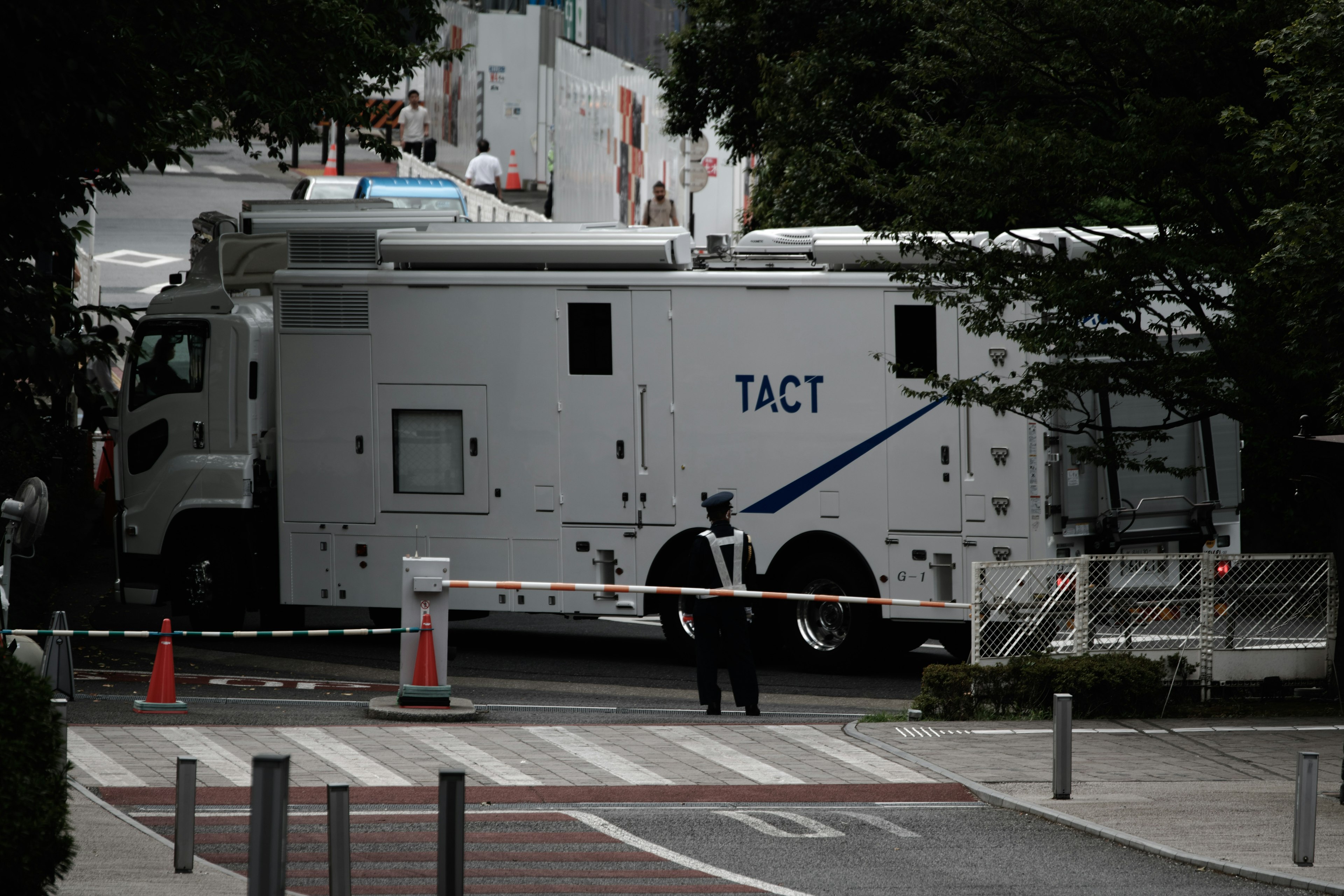 TACTと書かれた白い大型車両が見える通りの景色 警備員が周囲を見守っている