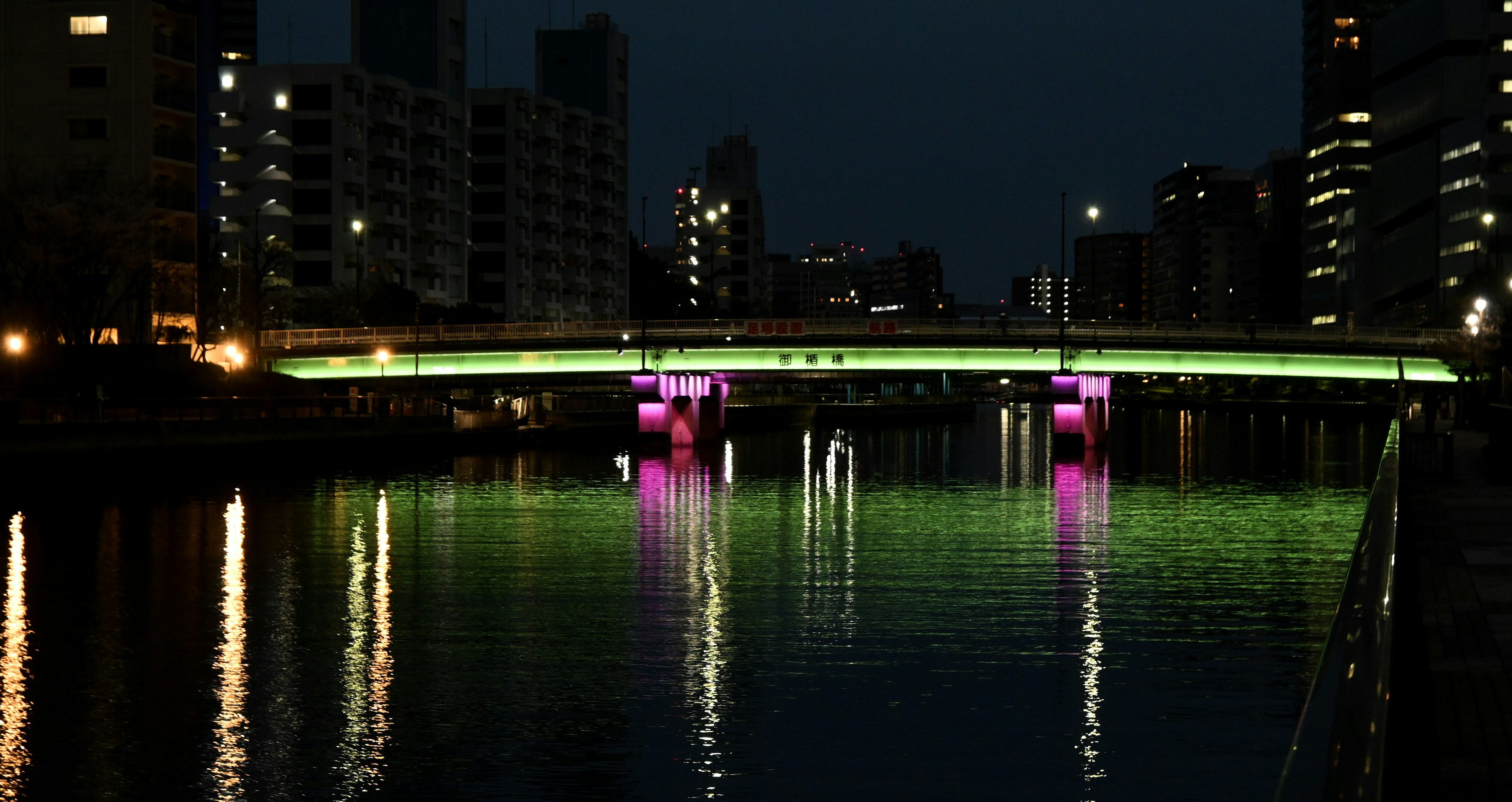 夜の都市景観に照らされた緑とピンクのライトが映る橋と水面