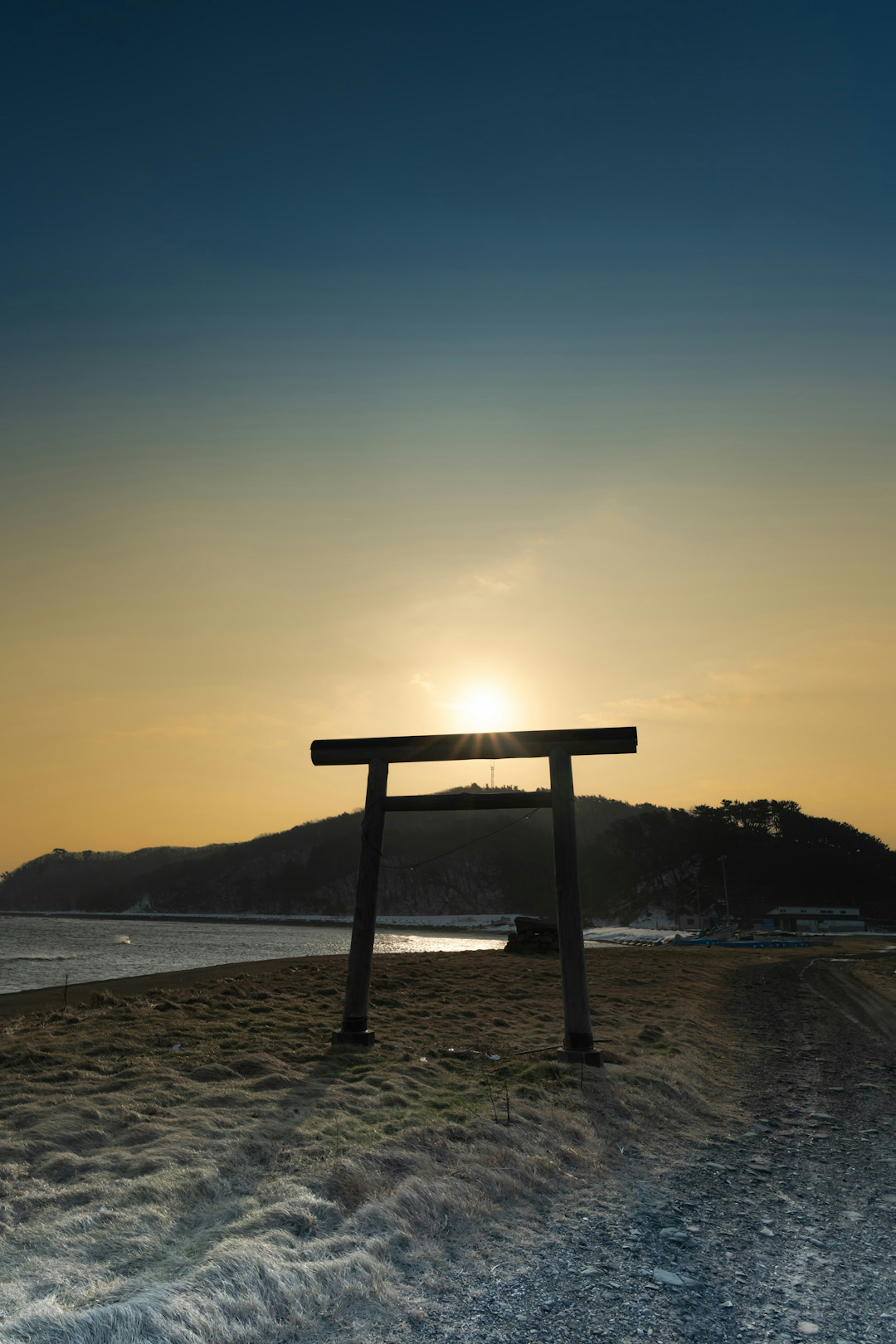 Gerbang torii yang disilhouetkan melawan matahari terbenam dengan pantai dan air