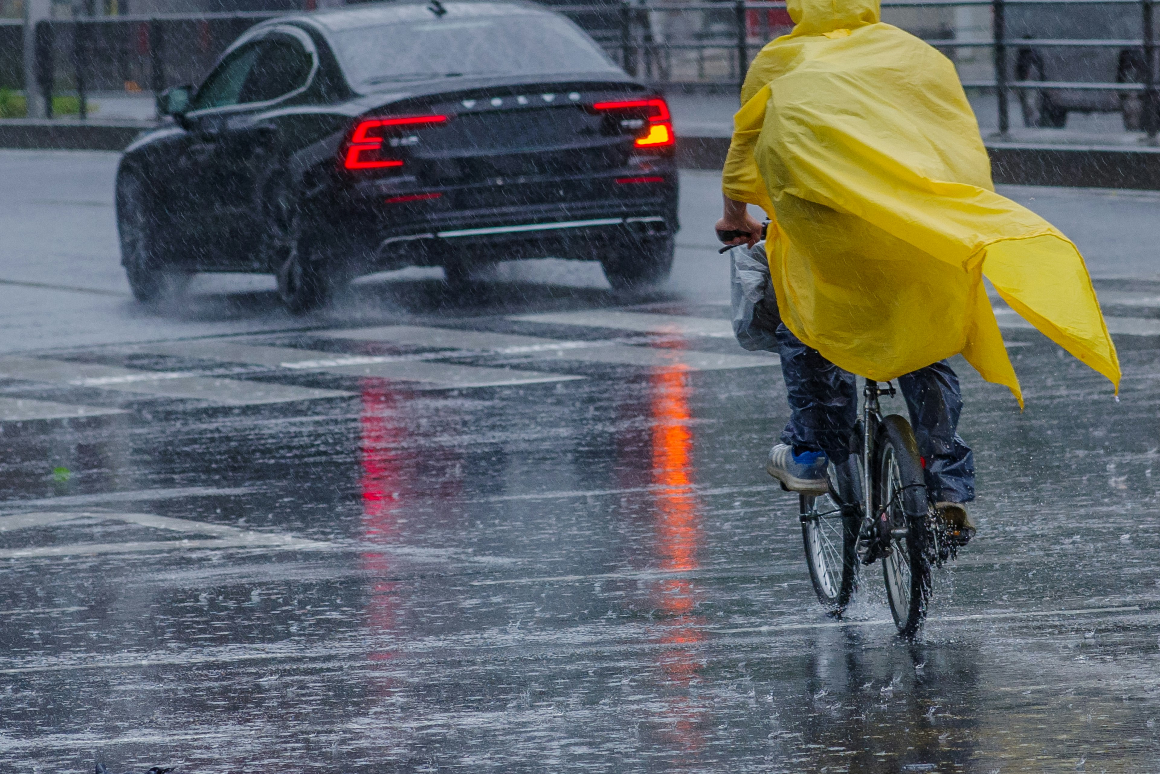 Une personne à vélo sous la pluie portant un imperméable jaune une voiture noire qui passe