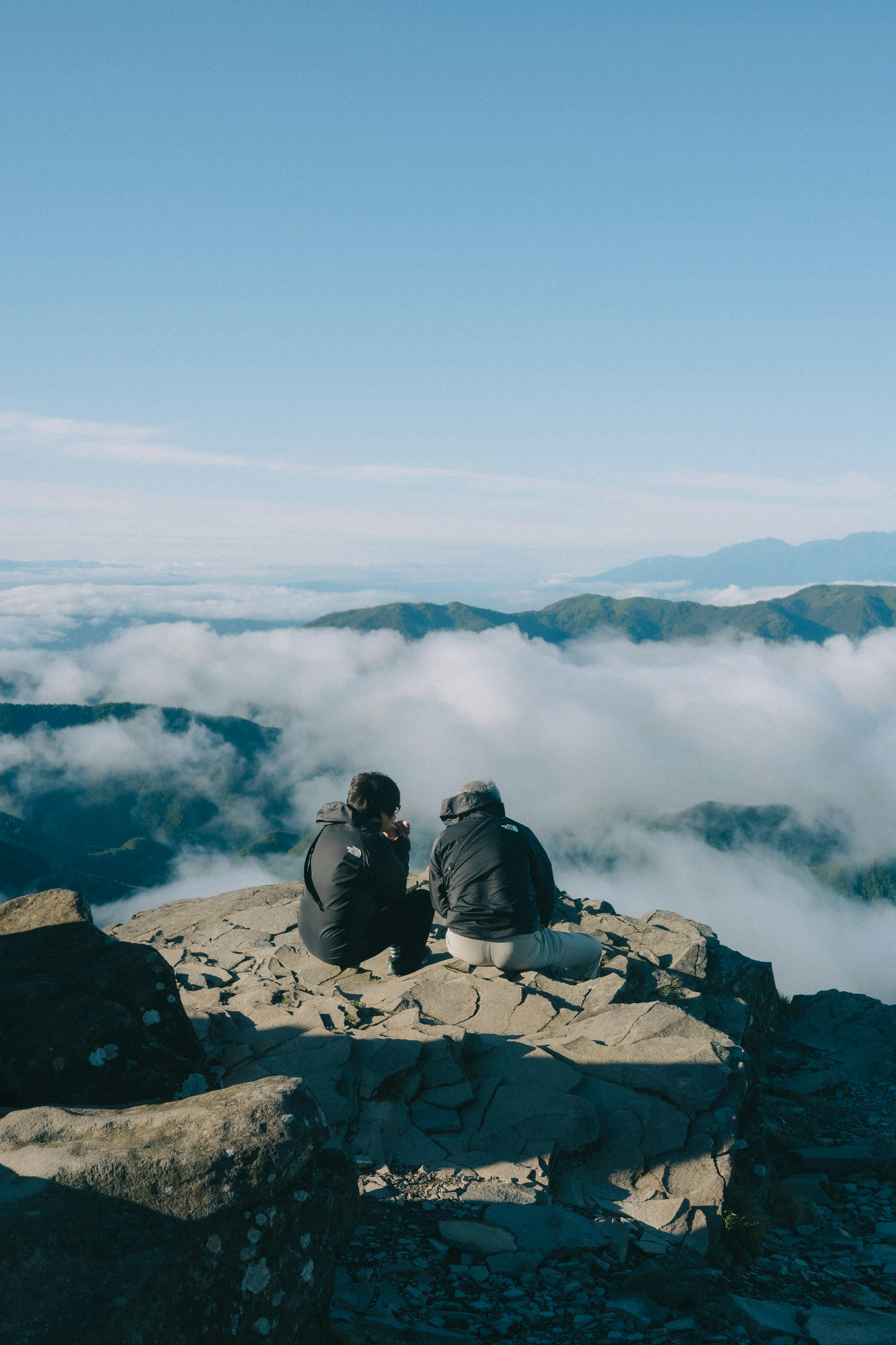 Zwei Personen sitzen auf einem Berggipfel und blicken auf ein Wolkenmeer