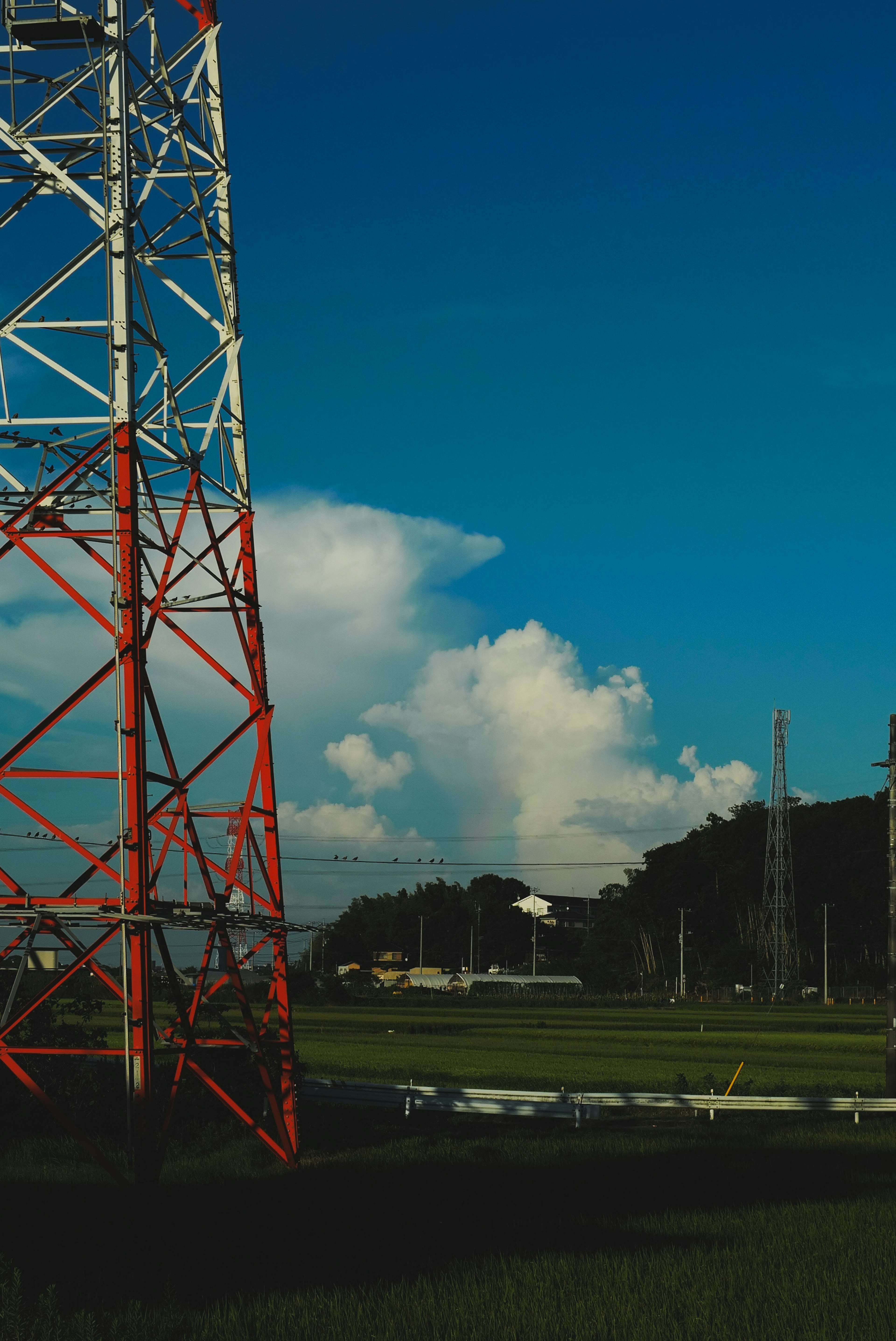 Menara komunikasi merah dan putih di bawah langit biru dengan awan dan lanskap hijau